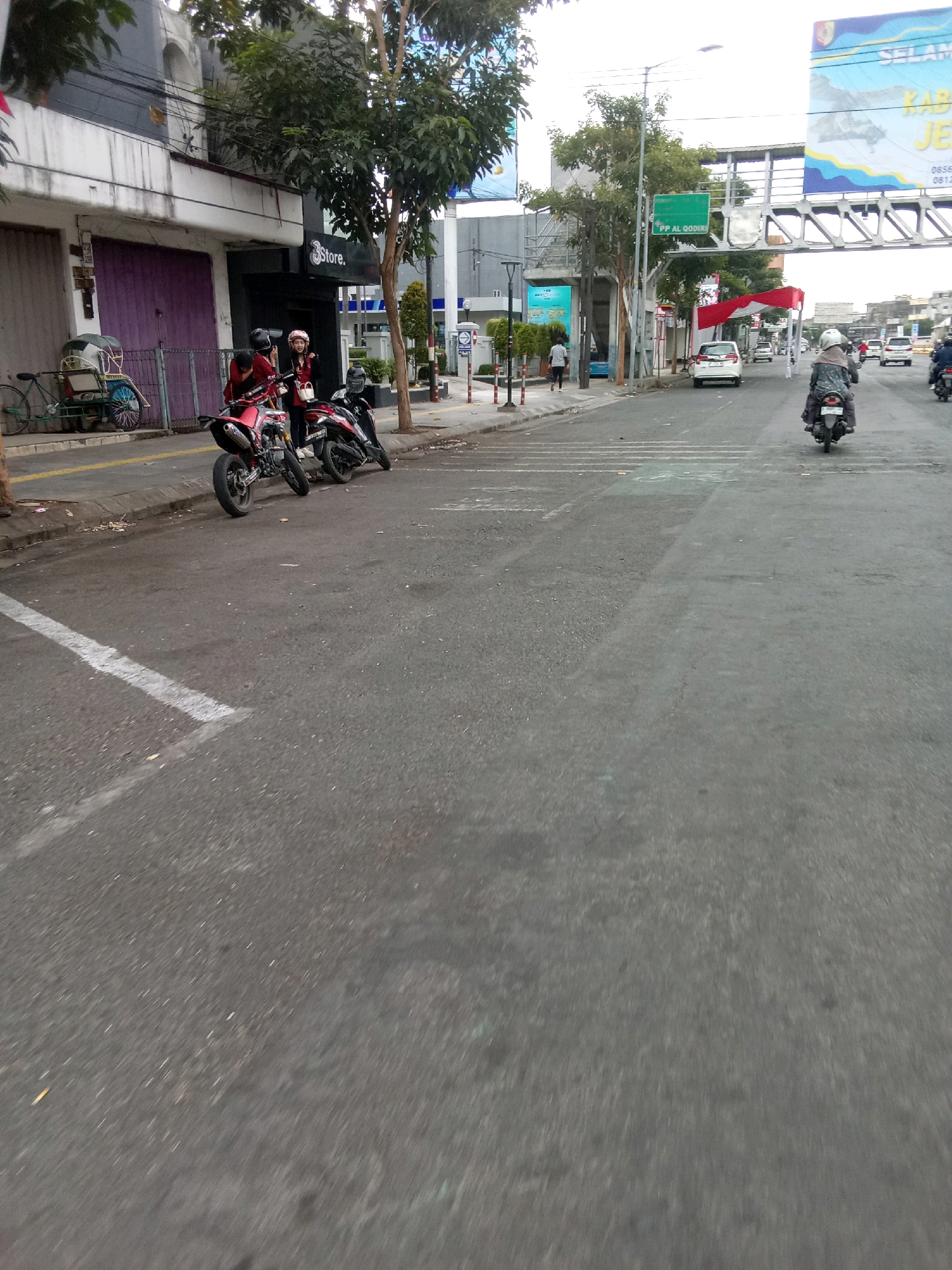 The image shows a street scene in Jember, Indonesia. Two people are on a red motorcycle on the left side of the street. Another person is on a black motorcycle further down the street. There are buildings on the left side of the street, including a shop with a sign that says "3store." There are also trees and streetlights. A large billboard is visible in the background. The road is paved and has white markings. The sky is overcast.