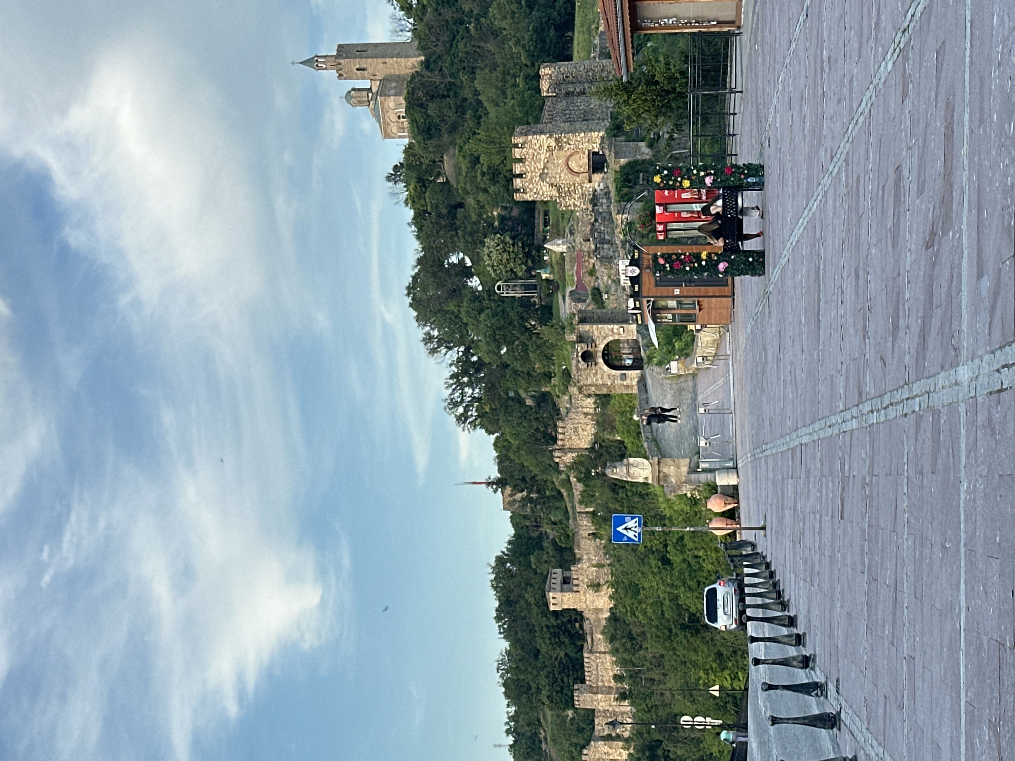 The image depicts a wide, stone-paved plaza in the foreground, lined with decorative black bollards on the left and a metal fence on the right. Two people are seated on a bench decorated with flowers, facing away from the camera, in the middle ground. Behind them, a small, wooden ticket booth is visible. Further back, a stone bridge or walkway leads up to a fortified wall with battlements, characteristic of Tsarevets Fortress in Veliko Tarnovo, Bulgaria. A pedestrian crossing sign is also present. The fortress walls and a church with a tower dominate the hilly landscape in the background, covered with dense green trees. A white car is parked on the left side of the plaza, and several large terracotta pots are placed near the bridge. The sky is blue with scattered white clouds.