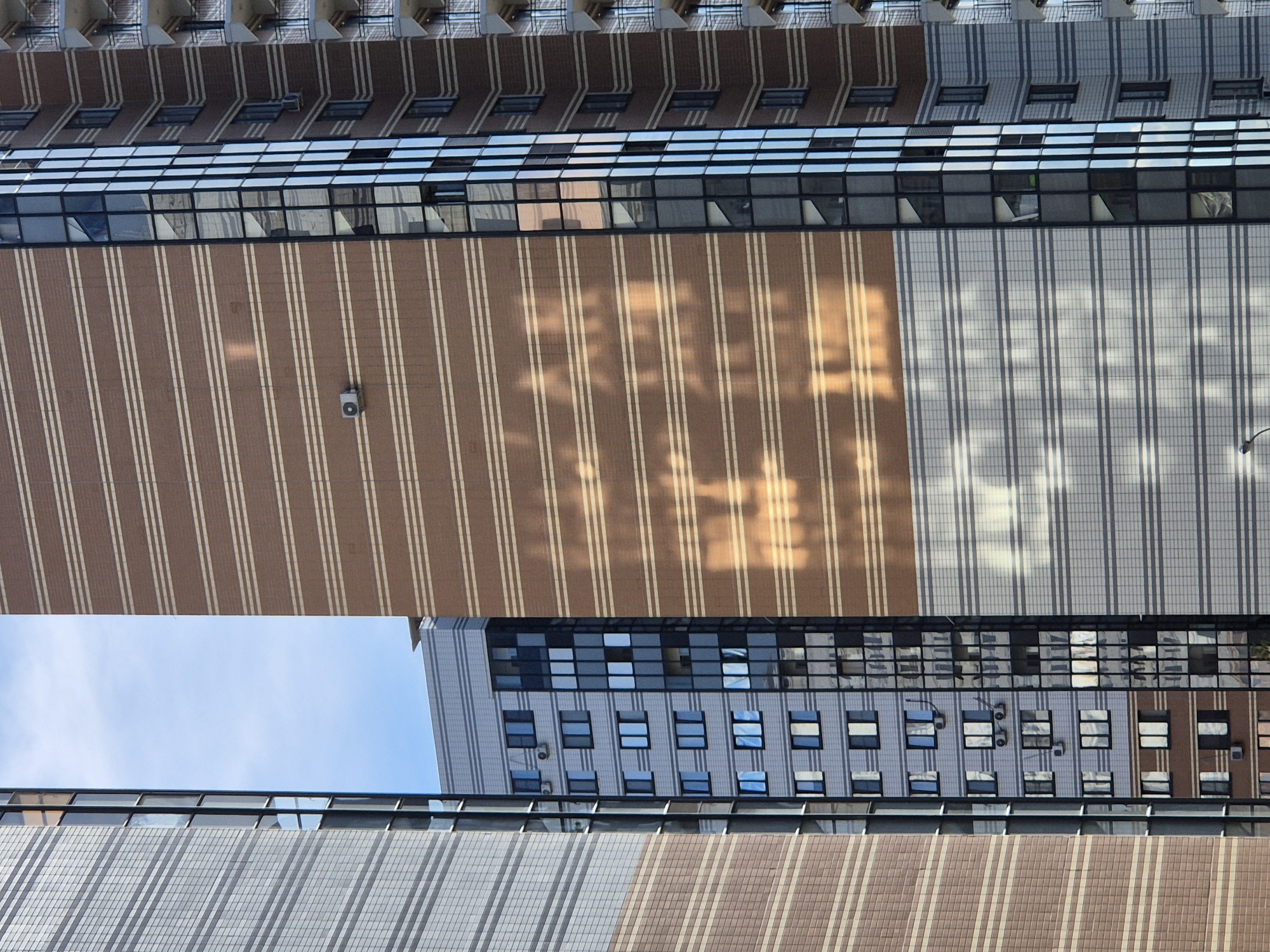 This image presents an abstract architectural perspective of modern buildings. The primary subjects are the building facades, characterized by repeating patterns of windows and textured surfaces. The architecture appears to be contemporary, featuring large expanses of glass and contrasting materials like brown brick and gray tiling.
The scene is an outdoor urban setting, likely a business district or a dense residential area, given the scale and style of the buildings. There are no people or specific objects of interest beyond the architectural elements themselves, except for a small camera or sensor mounted on the brown brick section.
The lighting suggests it is daytime, with sunlight casting distinct shadows and reflections on the building surfaces. The warm hues of the sunlight, particularly on the brown facade, create a visual interplay of light and shadow that forms abstract patterns. The sky is visible in a gap between two sections of the buildings, appearing clear and blue, indicating fair weather.
The image is taken from a high vantage point, looking down at an angle that emphasizes the geometric lines and repetitive modules of the architecture. The composition is dominated by vertical and horizontal lines, creating a sense of order and structure. The lack of discernible human activity and the focus on the building exteriors contribute to a feeling of impersonality and monumental scale.
There is no visible text in the image. The overall impression is one of urban grandeur and modern design, viewed through a lens of abstract pattern and light. Given the location context of Voronezh, Russia, this could be a view of an office complex, a university campus, or a large residential development.