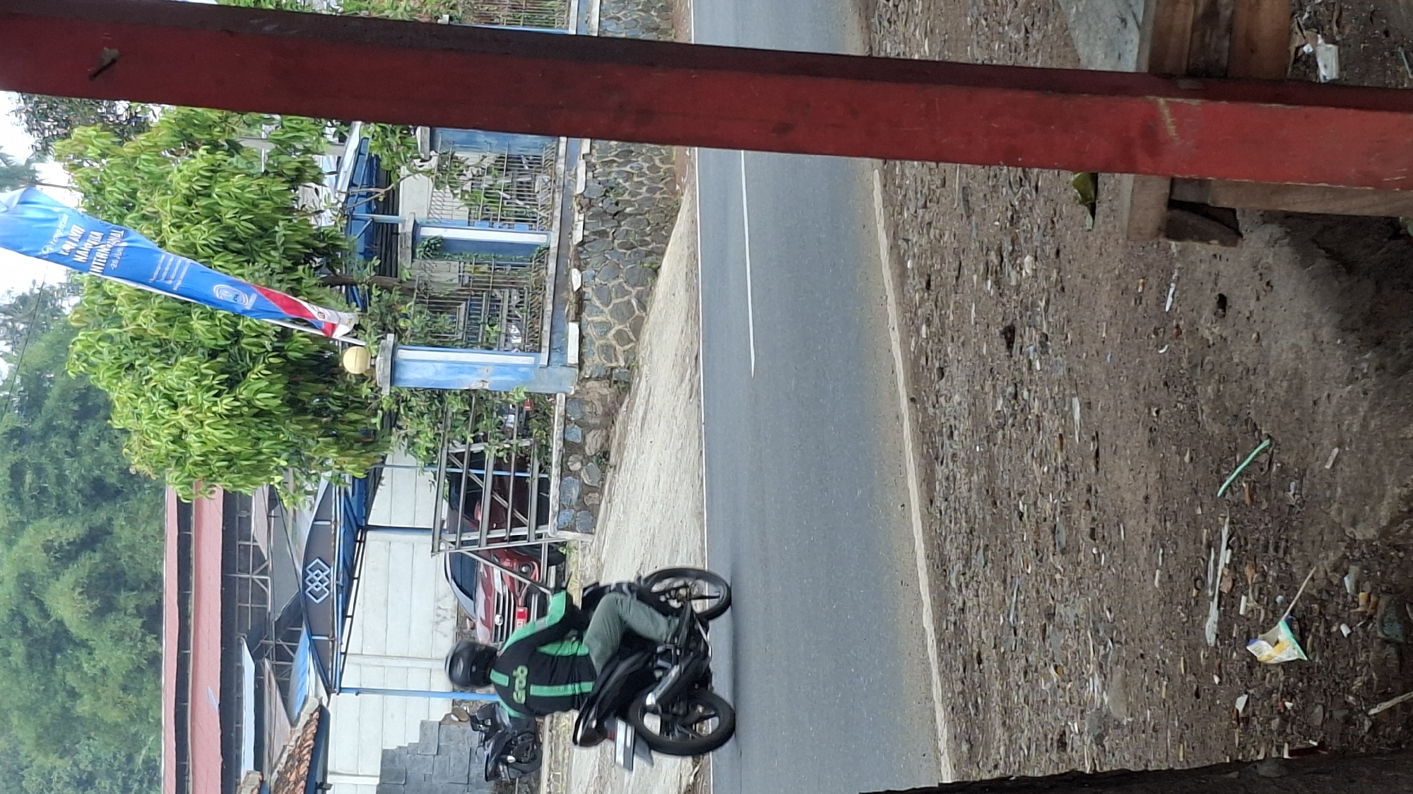 In Cilaku, Indonesia, a person wearing a black helmet and a green and black jacket with the "Grab" logo is riding a black motorcycle. They are on a paved road, with a small pile of trash on the side of the road. In the background, there are trees, a building with a red roof, and a blue fence. A blue flag with Indonesian writing is also visible. The overall scene is a street view on a cloudy day.