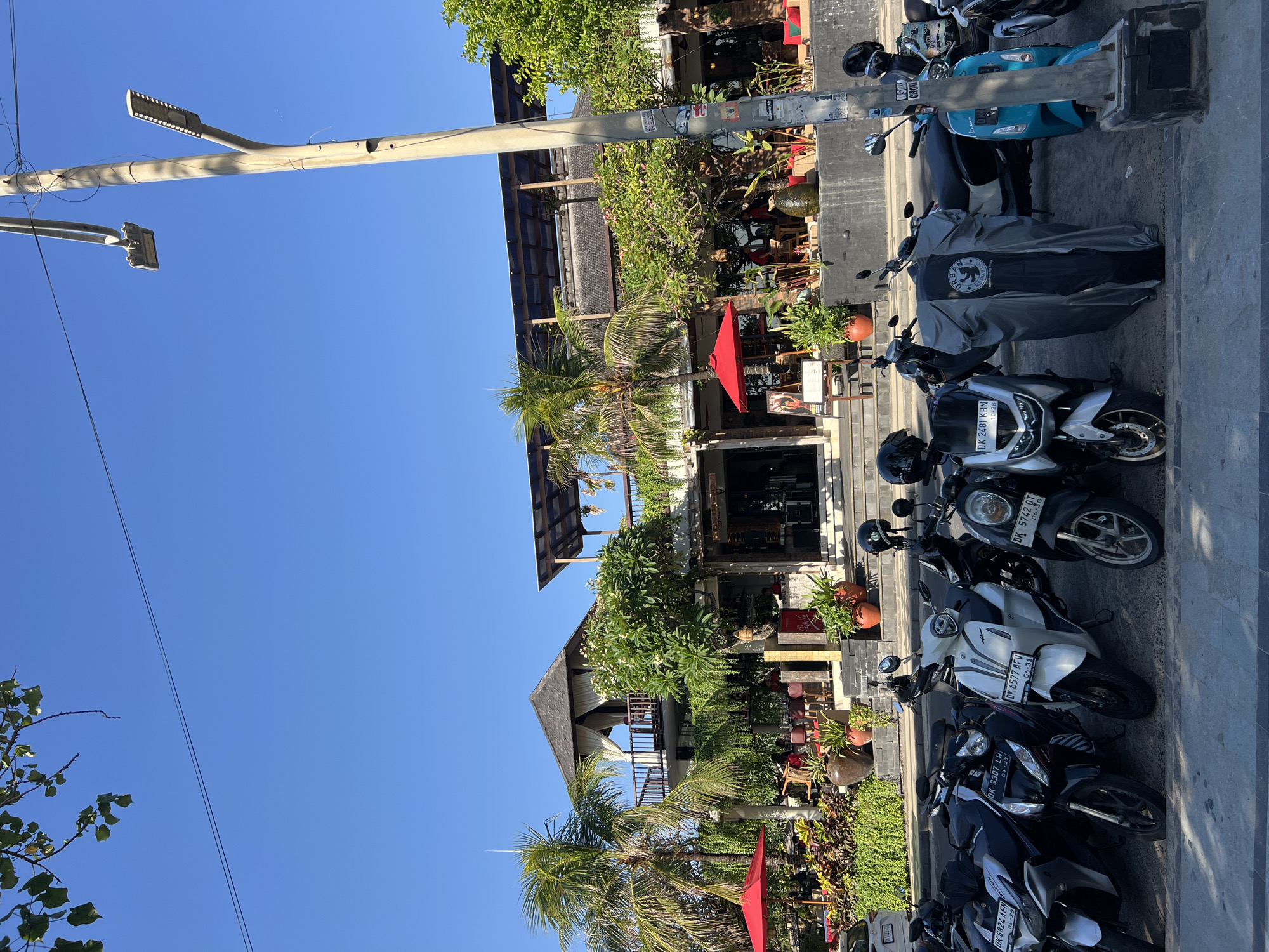 A wide, low-angle exterior shot captures a street scene in Kuta, Indonesia, on a clear, sunny day. The foreground is dominated by a row of parked motorbikes and scooters along a paved sidewalk. Among the vehicles, several have license plates visible, including "DK 6824 AEW," "DK 3307 LWH," "DK 6577 AFU," and "DK 5742 OT." One scooter is covered with a grey tarp bearing the "URBAN REPUBLIC" logo.
Behind the parked vehicles, a multi-story building stands, featuring a dark, sloped roof and an open-air upper level with wooden railings. Palm trees and lush green foliage surround the building, with two prominent red umbrellas visible at ground level. Planters with potted plants are arranged near the building's entrance. The building appears to be a restaurant or resort, with seating and awnings visible. A tall, grey utility pole with two streetlights stands on the right side of the frame, adorned with several stickers. The sky is a vibrant, clear blue.