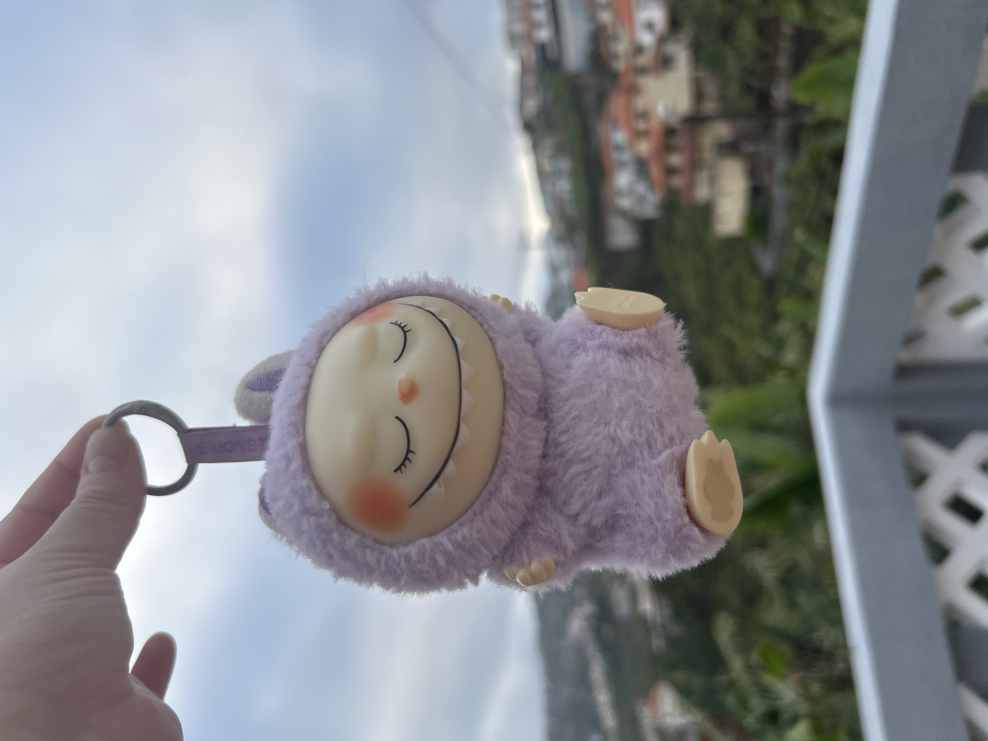 A close-up, slightly blurred, and rotated image shows a person's left hand holding a light purple, fuzzy sheep-like plush keychain with a round, plastic face featuring closed eyes, a smiling mouth, and blush on its cheeks. The keychain hangs from a metal ring connected to a fabric tab.
In the background, blurred structures and foliage indicate an urban setting. Through the railing, there is vegetation and the reddish-orange rooftops of buildings, consistent with the architecture found in the city of Câmara de Lobos, Portugal. The sky is visible in the top left of the shot. The image appears to have been taken from a slightly elevated position. A gray, grid-patterned railing is visible on the right side of the frame.