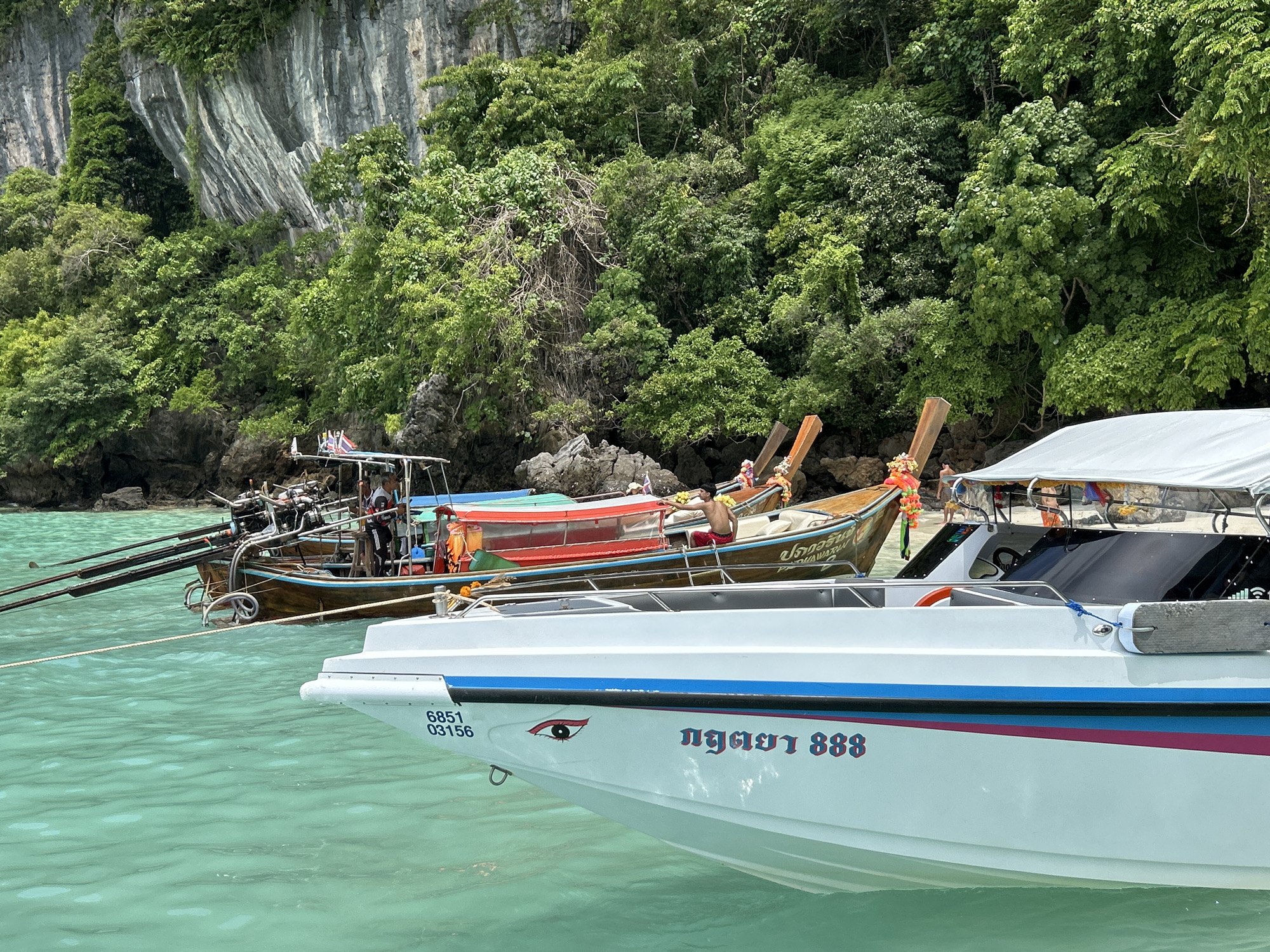 This image shows a tropical scene in Ko, Thailand, featuring several boats on turquoise water. In the foreground, the bow of a white speedboat with blue and pink stripes is visible. It has registration numbers "6851 03156" and a stylized eye decal on its hull. Thai script, reading "งูทะเล 888", is also present on the side of the speedboat.
Behind the speedboat, a traditional Thai longtail boat is moored. This boat has a wooden hull and is painted with bright colors, including red and orange. It is equipped with multiple longboat engines mounted at the stern. A Thai flag is visible on the longtail boat.
Further in the background, a sheer limestone cliff covered in dense green vegetation rises from the water. The water itself is clear and shallow, revealing hints of the seabed. The overall atmosphere is serene and indicative of a popular tourist destination.