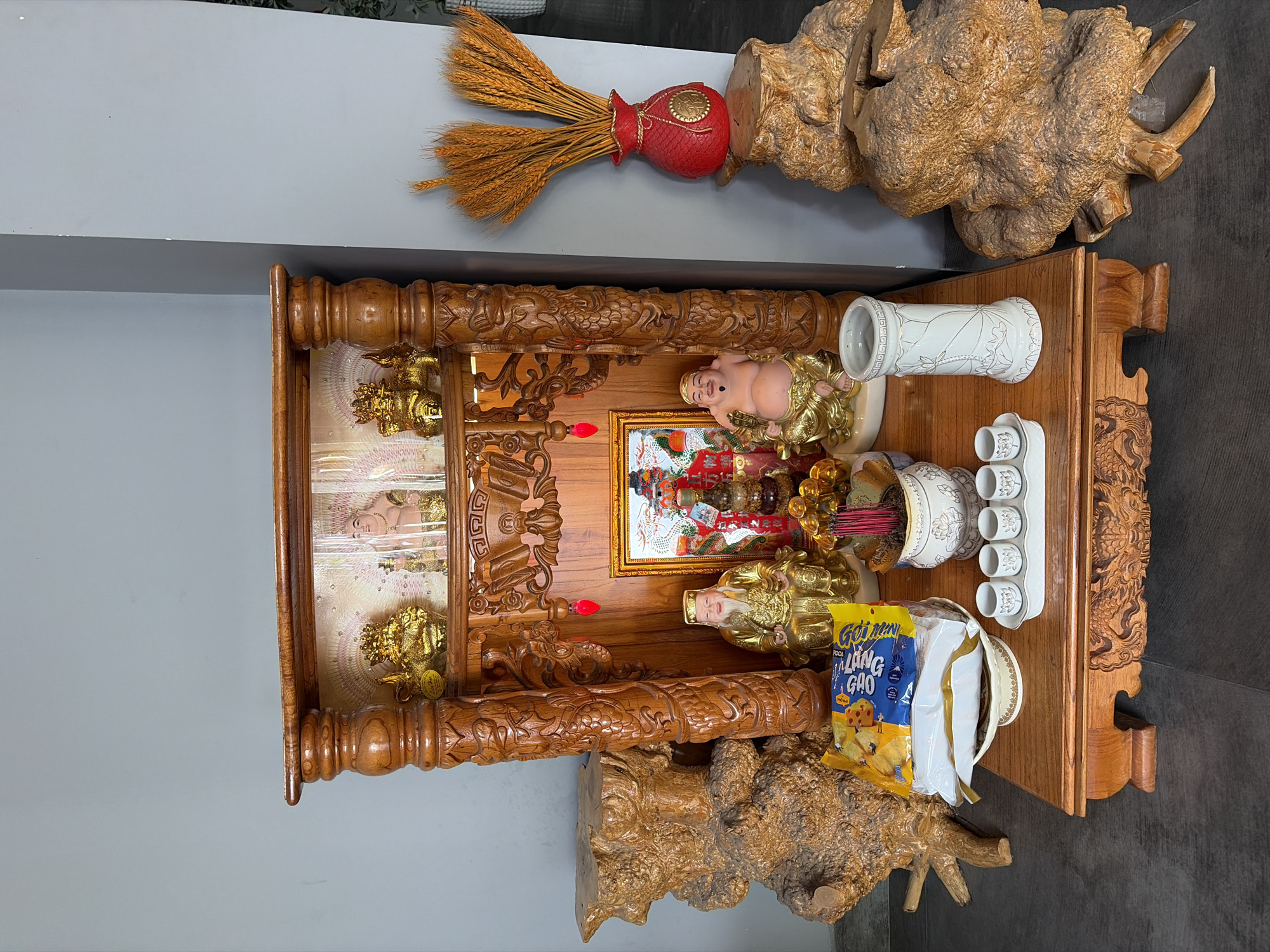 This is a medium shot, eye-level, full-frame photograph of a Buddhist altar. The altar is made of intricately carved dark wood with a glossy finish and is situated indoors, likely in a private residence in the city of Đà Nẵng, Vietnam.
Dominating the center is a tiered wooden structure, ornate with carved dragons and decorative motifs. The top tier features a framed image of a Buddha, flanked by two golden lion statues and a single golden figurine. The middle tier displays a framed picture of a celestial scene, possibly depicting deities or significant Buddhist figures, with a small red pendant hanging below. On either side of this central picture are two larger golden statues. The left statue depicts a seated man in golden robes with a long white beard, wearing a golden cap. The right statue is a corpulent, smiling figure, also in golden robes.
In front of the altar, a wooden table displays several items. A white ceramic bowl holding incense sticks is placed near the center. To the left of the incense bowl is a stack of items, including a bag of chips with Vietnamese writing on it, resting on a white and gold ceramic plate. To the right of the incense bowl is a white ceramic vase with a lotus design. In front of the vase is a white ceramic tray holding four small white teacups, also decorated with lotus motifs.
To the left and right of the altar are large, weathered pieces of dark, gnarled wood, resembling tree roots or stumps, that add a natural, rustic element to the scene. Behind the altar, to the right, a bright red vase shaped like a traditional Asian money bag holds a bouquet of dried wheat stalks. The background wall is a neutral, light gray color. The floor is covered with dark gray, textured tiles. The lighting is bright and even, suggesting an indoor setting with good artificial or natural light. There are no people visible in the image.