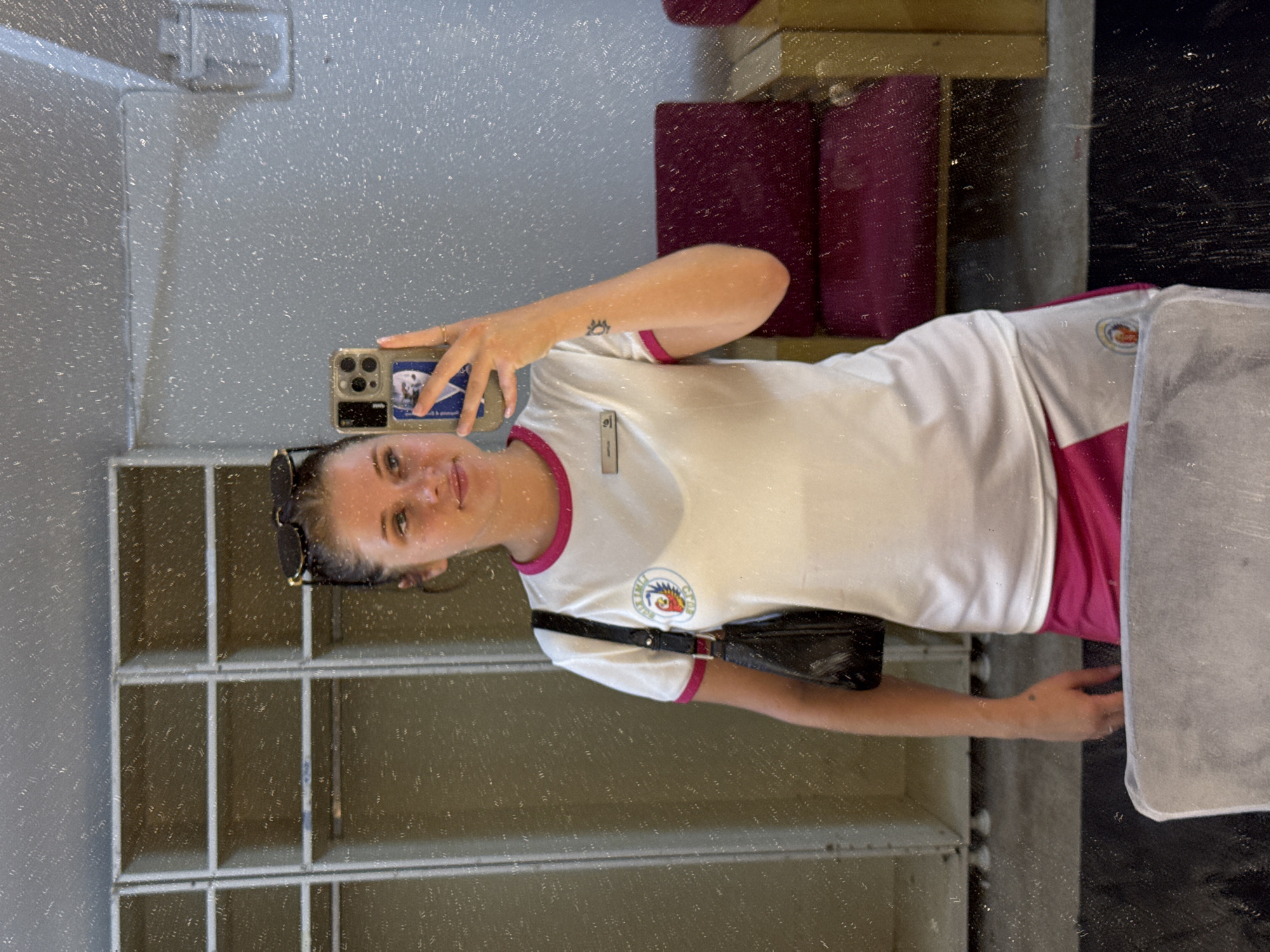 The image captures a young woman taking a selfie in a locker room environment, potentially in Aksu, Turkey. She is wearing a white t-shirt with pink trim and a matching pink skirt. Her hair is pulled back, and she has sunglasses on her head. She is holding a smartphone, which displays a sticker of a person's face. A black purse with a strap is slung over her shoulder. In the background, metal lockers are visible, as well as a burgundy chair. The mirror in which the selfie is taken is speckled with what appears to be dirt or dust, creating a textured effect on the image. A tattoo of a sun-like symbol is visible on her left forearm.
