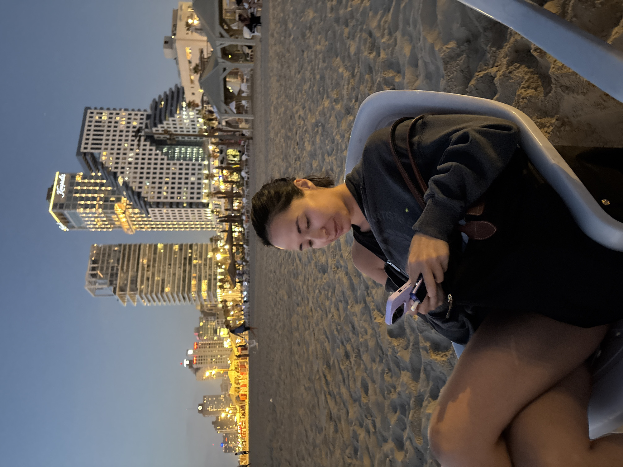 A woman sits in a white plastic chair on a sandy beach in Tel Aviv, Israel, looking down at her phone. The sky is a dusky blue, indicating dusk or twilight. In the background, the cityscape of Tel Aviv is visible, with numerous tall buildings illuminated by lights. Some of these buildings appear to be hotels or high-rise apartments. Along the beachfront, there are several white canopy structures and rows of chairs, suggesting a beach club or resort area. People can be seen walking and sitting in the distance. The woman is wearing a dark gray hoodie and black shorts, with a brown purse strap visible over her shoulder. Her phone is purple. The sand is textured and uneven.