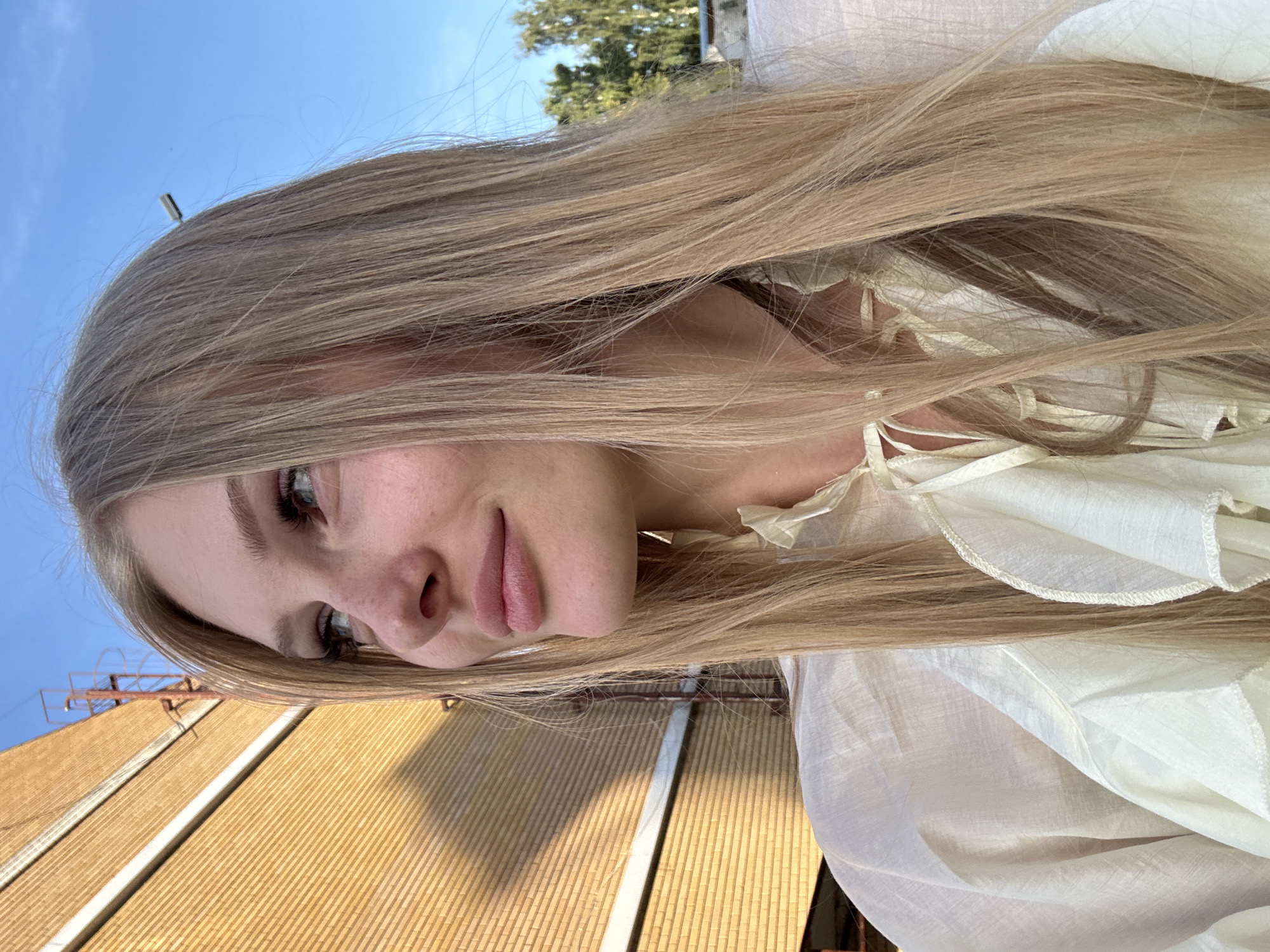 An outdoor, medium shot captures a young Caucasian woman from the chest up, facing right and looking upwards with a slight smile. She has long, straight, blonde hair with a subtle ombre effect, falling around her shoulders. Her eyes are blue, and she has a smattering of freckles across her nose and cheeks. She is wearing a loose, cream-colored blouse with ruffled details around the neckline and chest, tied with fabric strings. The background consists of a textured, yellow brick building on the left, with a shadow cast across it. Above and behind her, a clear blue sky is visible, with some distant green trees and structures on the right. The lighting suggests it is daytime, possibly late afternoon, with sunlight illuminating the scene. No specific landmarks in Kazan, Russia, can be identified from the visual information.