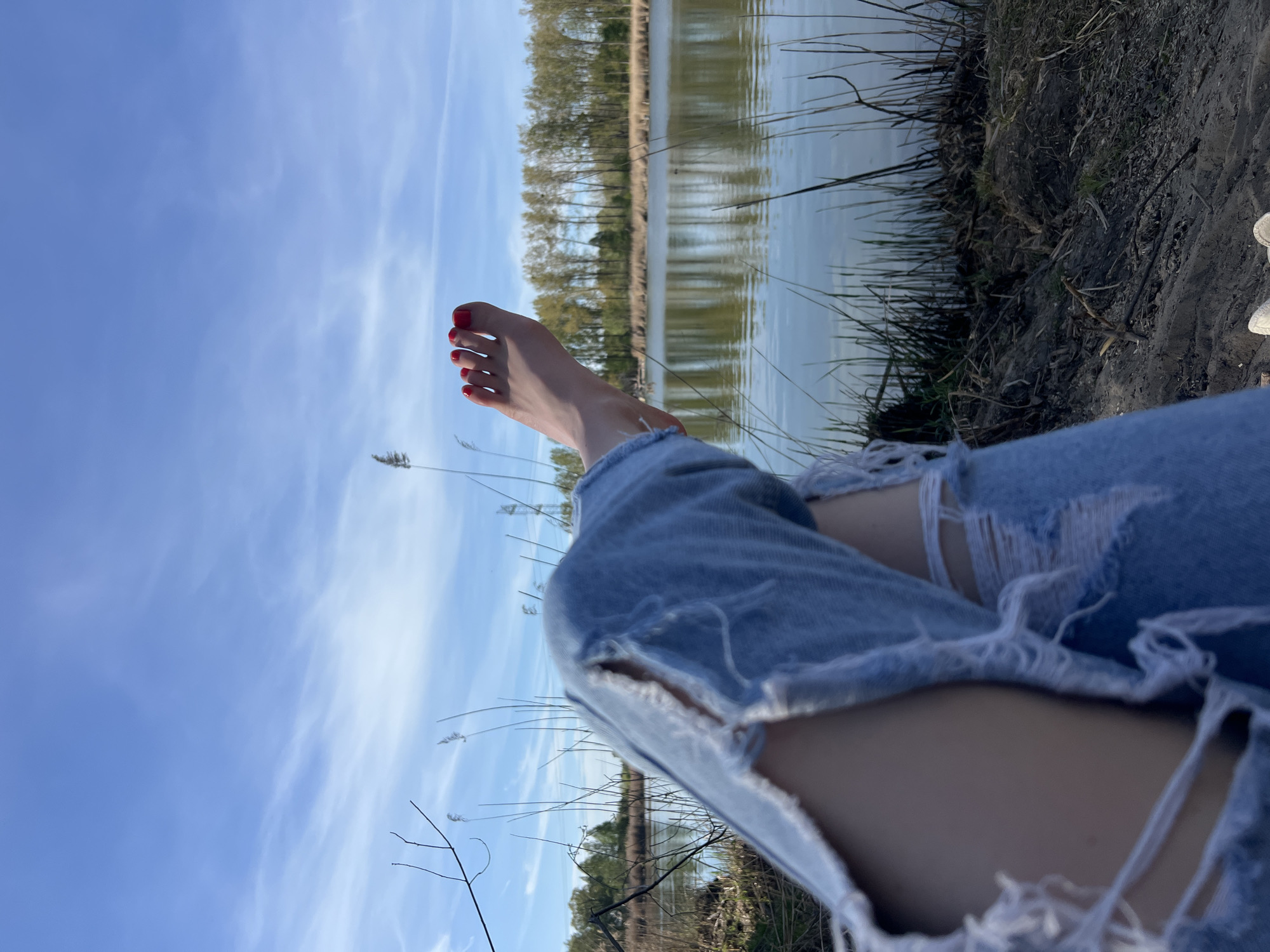 A person wearing ripped blue jeans sits at the edge of a body of water near Ciemne, Poland. Their bare foot, with red nail polish, is extended upwards toward the sky. The background includes a lake or river bordered by trees and vegetation. The sky is visible with scattered clouds. The foreground includes dry grass and other vegetation along the shoreline.