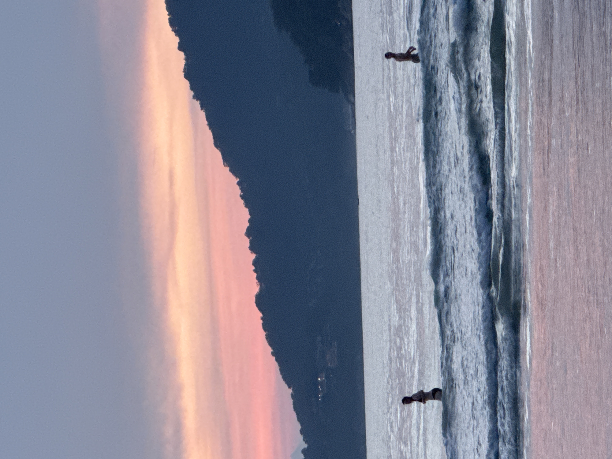 A wide shot captures two individuals wading in the shallow waters of Patong Beach, Thailand, during twilight. The sky displays a gradient of soft blues and pinks, with a dusky hue settling over a dark, tree-lined hillside in the background, dotted with distant lights. The sea is calm, with gentle waves lapping the shore. The foreground is dominated by the ocean, with the pinkish tones of the wet sand visible at the very bottom of the frame. The left figure, a person wearing a dark bikini, stands facing away from the viewer. The right figure, a person in what appears to be swimwear, is also facing away, further out in the water. Both figures are silhouetted against the shimmering sea.
