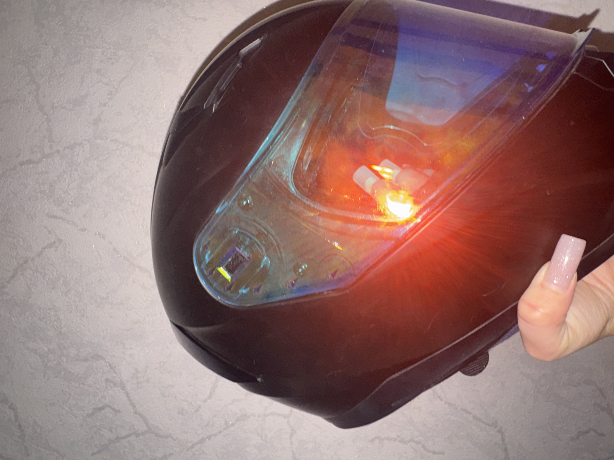A dark brown motorcycle helmet is held by a hand with long, light pink, glittery nails. The helmet has a reflective visor with blue and purple hues. A bright orange light is reflected on the visor, creating a lens flare effect. The background is a textured, light grey wall. The image is likely taken indoors, possibly in a residential setting in Bishkek, Kyrgyzstan, given the context. No other people or significant environmental details are discernible.