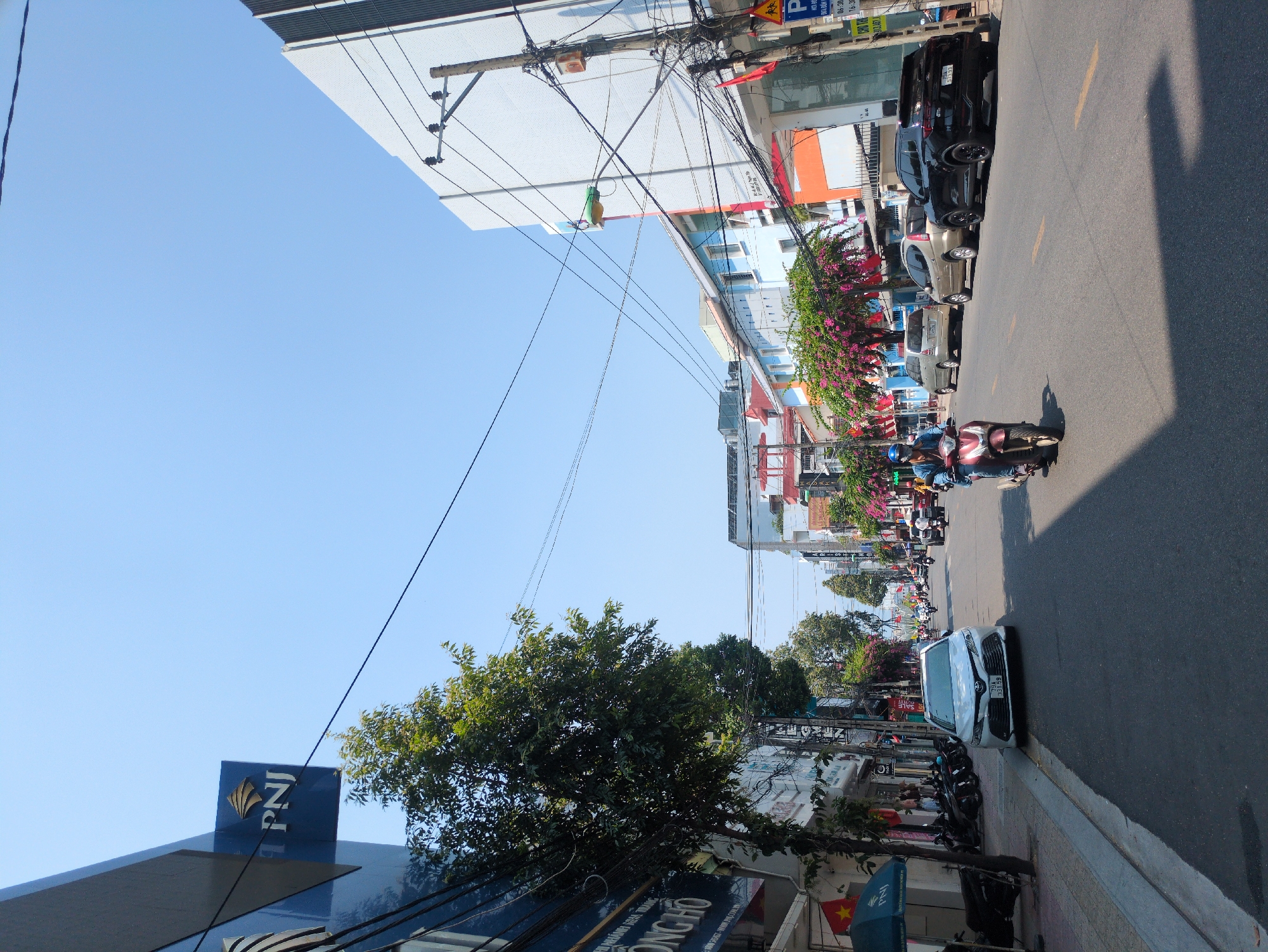 The image shows a street in Nha Trang, Vietnam. The view is along a busy street with buildings on both sides. Several parked cars line the right side of the street, along with a number of parked motorbikes. A white Toyota Camry is prominent on the right-hand side.
Traffic, primarily consisting of motorbikes, moves along the road. At least one motorbike is visible with two riders.
The buildings are a mix of commercial and residential, some with foliage spilling over the balconies. Power lines are strung haphazardly above the street.
The sky is clear and blue. The light indicates that it is likely midday, and shadows are cast on the street.
