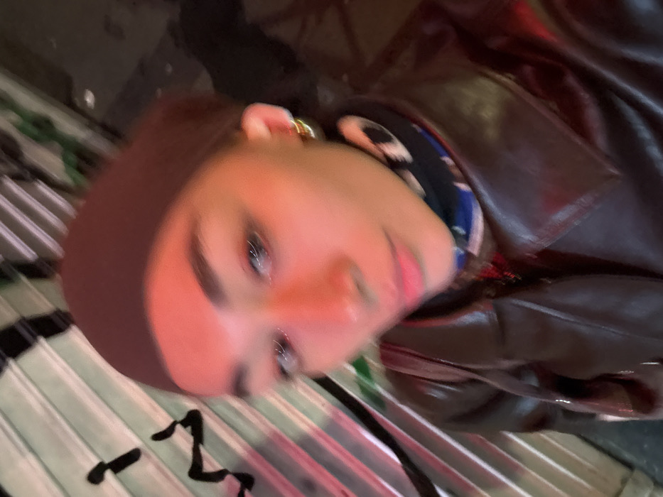 The image displays a close-up, diagonally tilted shot of a person's face and upper torso. The person has warm-toned skin, illuminated by a soft, warm light that casts a reddish-pinkish glow across their cheeks and nose. Their eyes are open and looking towards the camera, reflecting light, with dark eyebrows and visible eyelashes. A pinkish tint is visible on their lips.
They are wearing a dark brown head covering, which frames their face, and a dark brown jacket or coat, possibly made of leather or faux leather, with visible lapels and collar details. A patterned garment, featuring elements of blue, black, and white, is visible underneath the jacket around the neck area. A small, gold-colored earring is present on their visible earlobe.
The background is mostly dark and blurred, suggesting an indoor or urban setting at night. To the lower left of the frame, a distinct surface with horizontal parallel lines is visible, possibly a corrugated panel or a similar textured material. On this surface, there are abstract, black squiggly marks or lines. The overall impression is a casually posed shot, likely a selfie, with a slightly soft-focus effect.