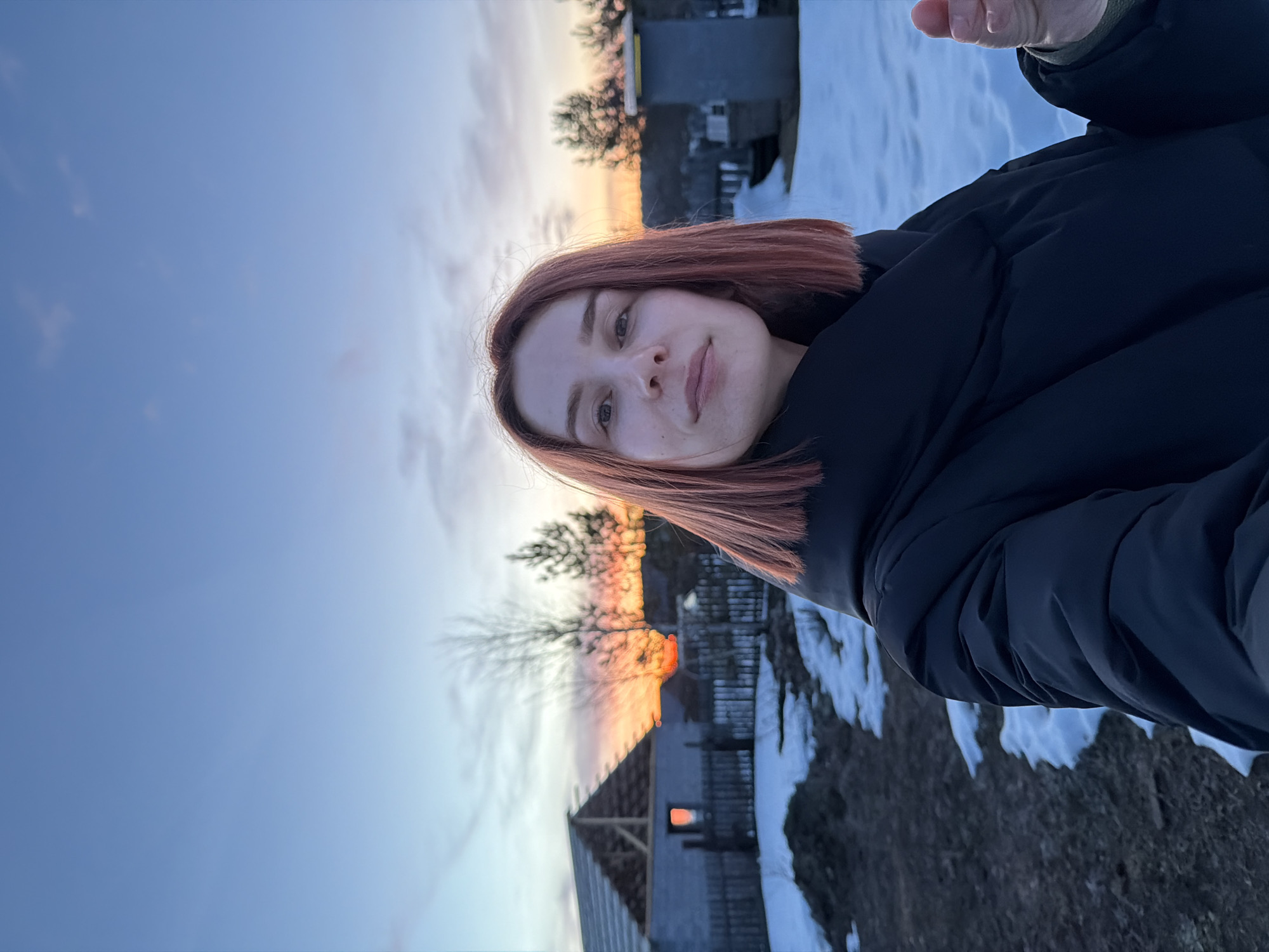 The image shows a selfie taken by a young woman with reddish-brown hair. She is wearing a black puffer jacket. The background suggests a rural or semi-rural setting in Chaikovsky, Russia, with patches of snow on the ground and a partially constructed wooden building visible. The sky displays a sunset, with orange hues near the horizon. Trees and what appears to be a fence are silhouetted against the light.