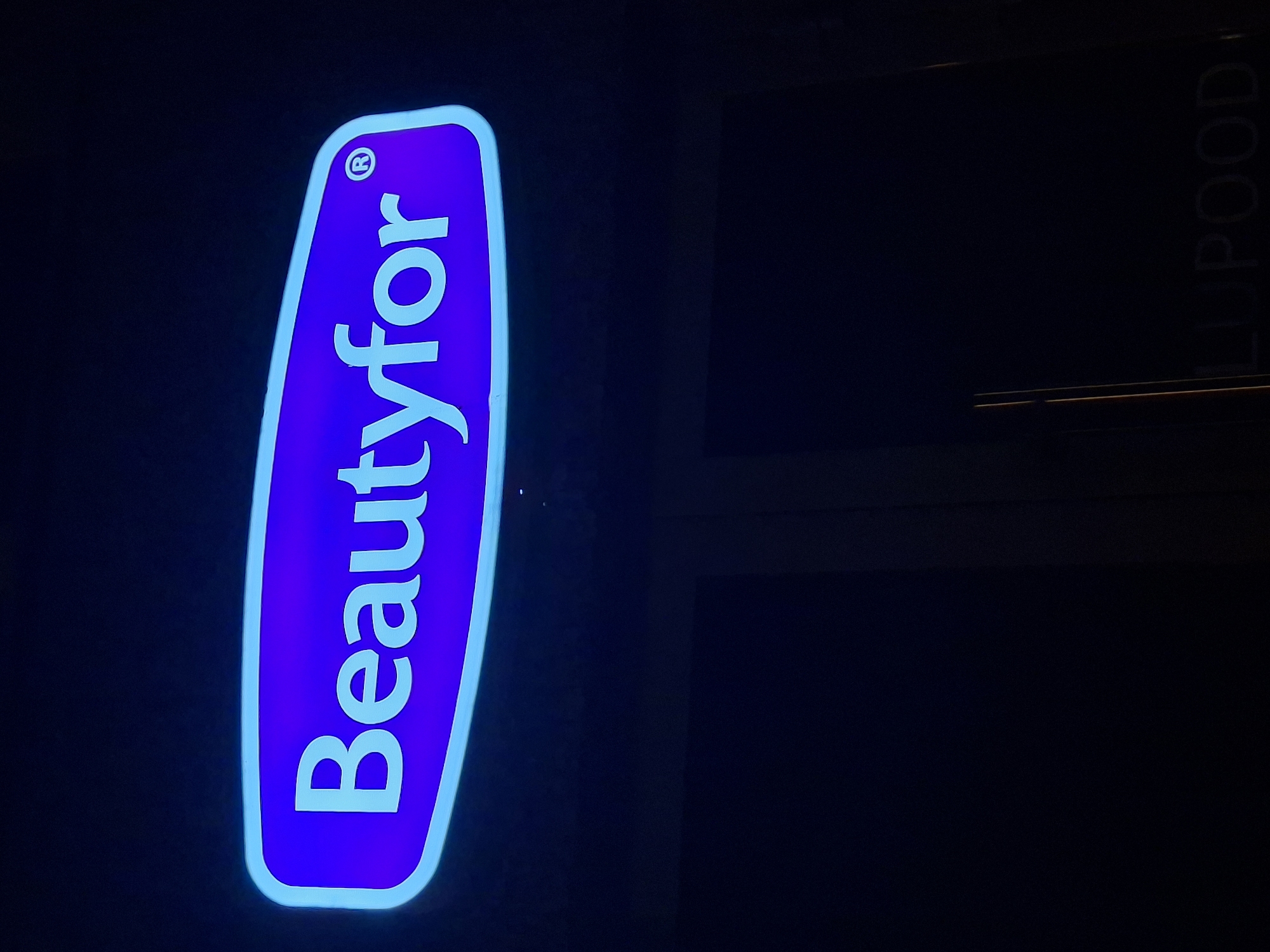 The image shows a neon sign that says "Beautyfor" in white letters on a bright purple background, outlined in a lighter blue. A registered trademark symbol "R" is visible on the right side of the sign. The sign is illuminated by neon lights and is likely mounted on the exterior of a building at night. In the lower part of the image, a dark area suggests a street or doorway, with faint vertical lines possibly indicating architectural features or more signage. A dimly lit sign with the text "LUPOOD" is partially visible at the bottom. The overall setting is dark, consistent with nighttime in an urban environment. Based on the visible text and common knowledge of businesses in Tallinn, "Beautyfor" is a business located in Tallinn, Estonia.