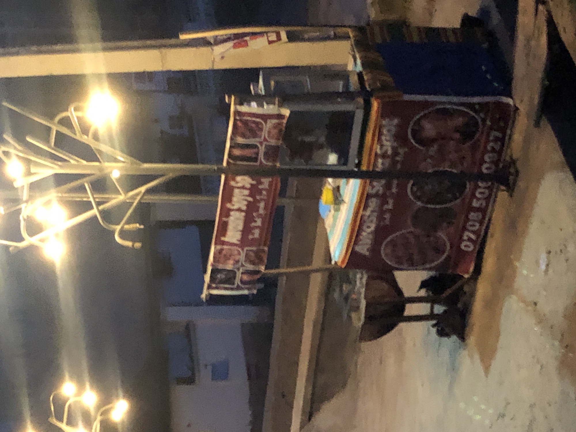 The image displays an outdoor scene at night, featuring a food vending stall and a distinctive street light. The setting is dark, with artificial lights providing illumination.
A prominent street light stands on the left, characterized by a central pole from which several decorative, multi-pronged arms extend, each capped with a bright, glowing bulb. The light casts a warm, intense glow that partially overexposes parts of the image.
In the center-right, a food stall or cart is set up. It features two large red banners. The banner on the left, partially obscured by the street light, clearly shows the text "Awesome Suya Spo...", likely indicating "Awesome Suya Spot". Below this text are smaller square images depicting various food items. The banner on the right, which is more visible, displays "Awasha Suya Spot" and a phone number: "0708 500 0827". This banner also features several circular images of what appear to be different types of suya (grilled meat skewers). The stall has a multi-colored striped canopy over its counter.
Underneath the stall, in the deep shadows, a small, dark shape, possibly an animal like a cat or dog, can be vaguely discerned. The ground around the stall is uneven, showing textures of pavement or dirt, reflecting some of the light. In the distant background, blurred and dark structures suggest residential or commercial buildings. The overall scene depicts a typical night-time street food vendor operating under artificial light.