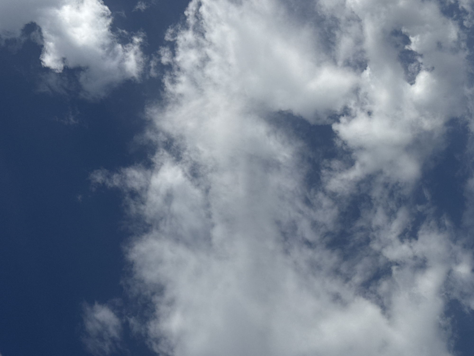 The image is a full-frame, high-angle shot of a deep blue sky with scattered white clouds. The clouds vary in density and texture, with some appearing wispy and others more opaque and billowy. The lighting suggests it is daytime, with the sun likely positioned above or behind the viewpoint, illuminating the upper surfaces of the clouds. There are no visible landmarks, people, or objects that can be used to determine a specific location within Sary-Oy, Kyrgyzstan. The scene is purely natural, depicting a sky on what appears to be a clear to partly cloudy day.