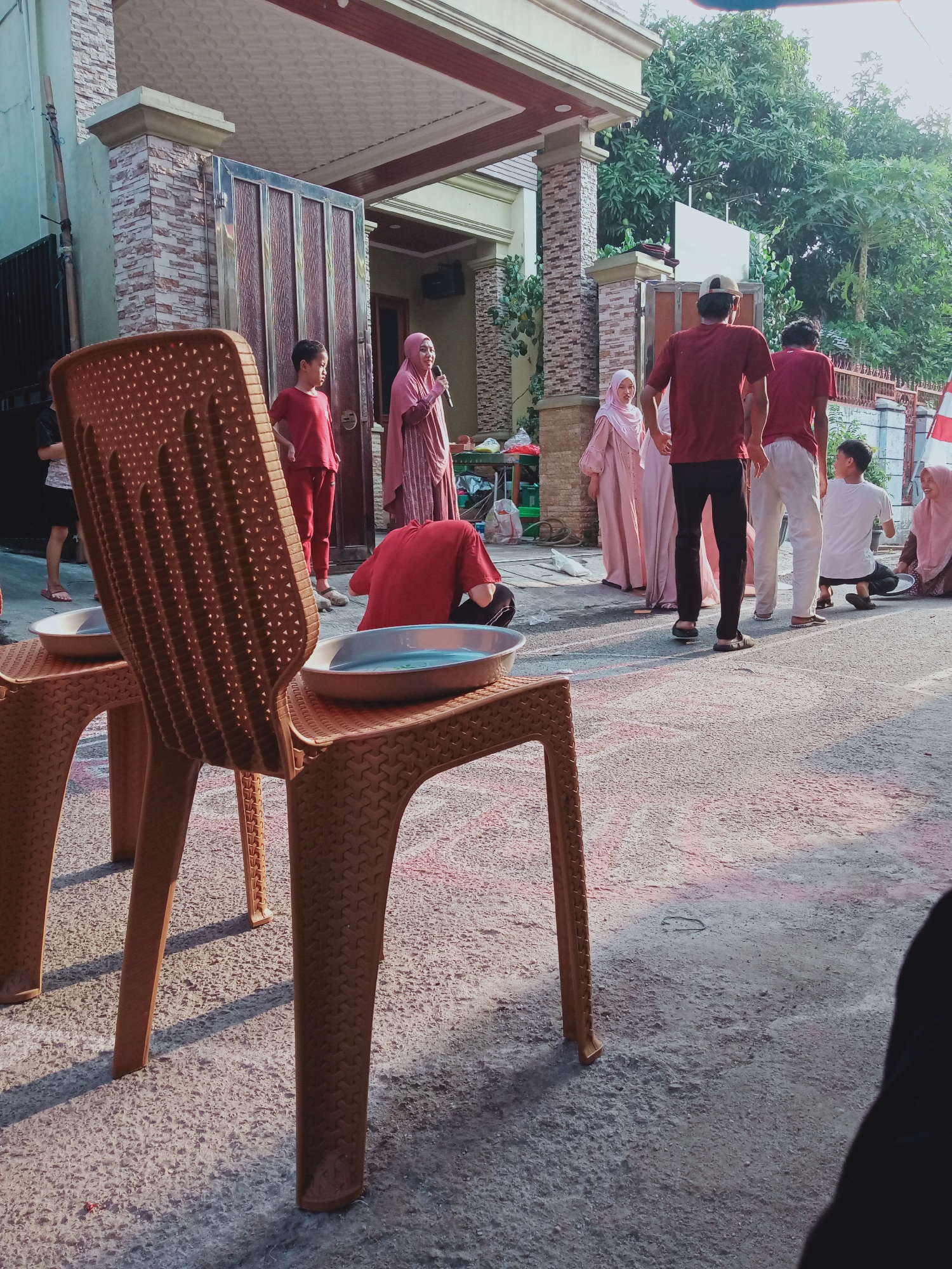 The image depicts an outdoor scene, possibly a residential area in East Jakarta, Indonesia, with several people gathered. In the foreground, two brown plastic chairs are positioned. One chair is mostly visible, showing a patterned texture and a shallow blue basin on top. The second chair, to the left, is partially obscured.
A group of individuals are standing and sitting in the background. One woman in a pink hijab stands near a large metal gate, holding a microphone, seemingly addressing others. To her right, a boy in a red shirt and pants stands. Further to the right, three individuals in maroon shirts and white pants are standing, with two facing away from the camera. Another person in a white shirt and dark pants is seated. To the far right, a woman in a pink hijab is seated on the ground.
The environment includes a building with a stone-patterned facade and a large metal gate. Trees and greenery are visible in the background. The ground is asphalt, with some red and pink markings, suggesting a game or activity might have taken place. The lighting indicates it is daytime, with shadows cast by the chairs and people.