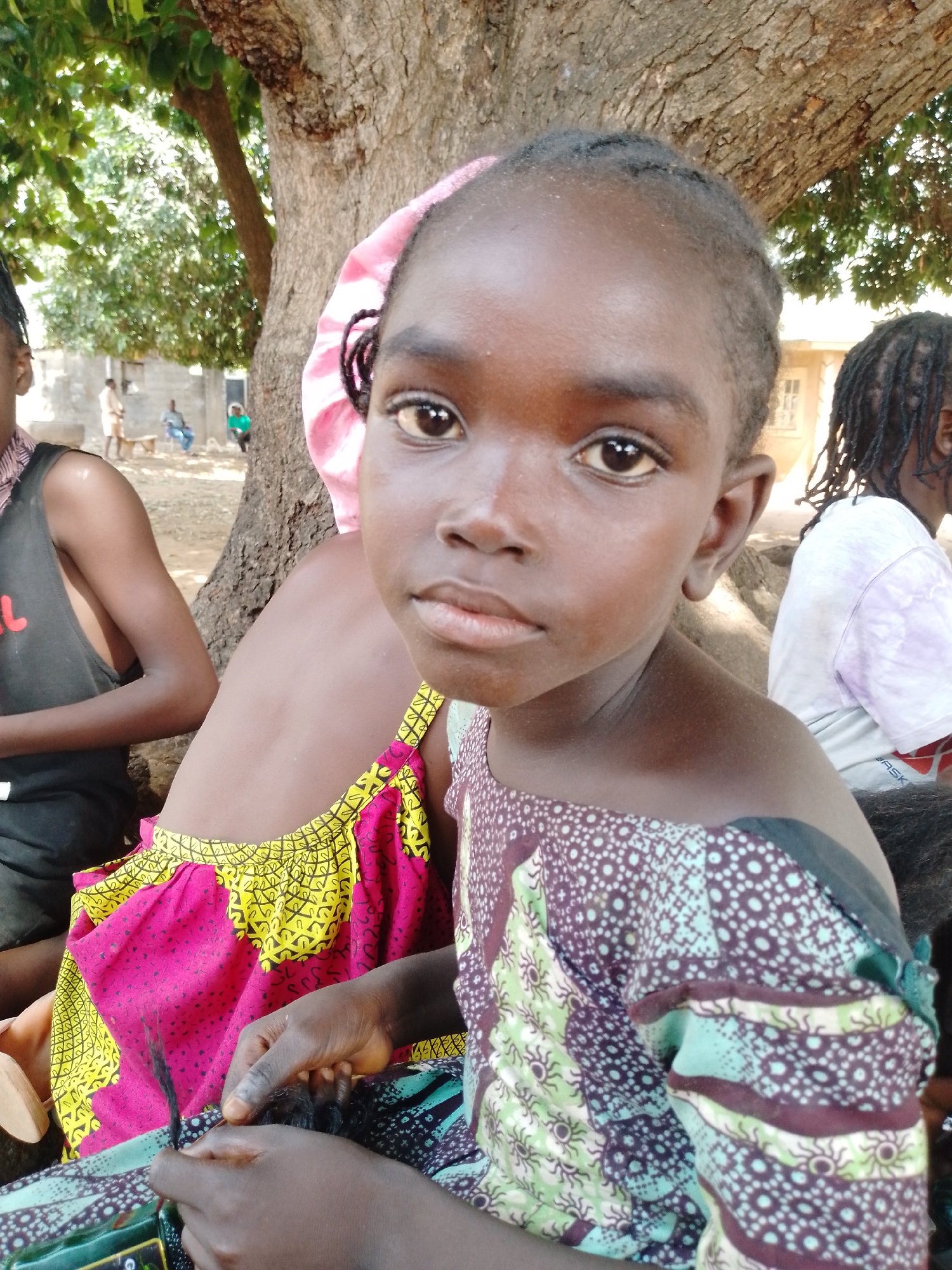 A young girl is the focus of this outdoor photograph taken in an indeterminate location within the city of Unknown, Unknown. She has dark skin, short hair styled with small braids, and a neutral expression on her face. She is wearing a patterned dress with a combination of green, brown, and white colors.
Adjacent to her, partially visible, is a person wearing a brightly colored dress with pink and yellow patterns. This individual has a pink head covering. To the left of the girl, another person is present, wearing a sleeveless black shirt, their body partially cropped from the frame. In the background, further to the left, other individuals are visible sitting and standing in front of a light-colored building. Another person is seated to the right of the young girl, partially obscured.
The group is situated near the base of a large tree, with its trunk and branches visible above and behind them, providing shade. The overall scene is bathed in natural sunlight, suggesting it is daytime.