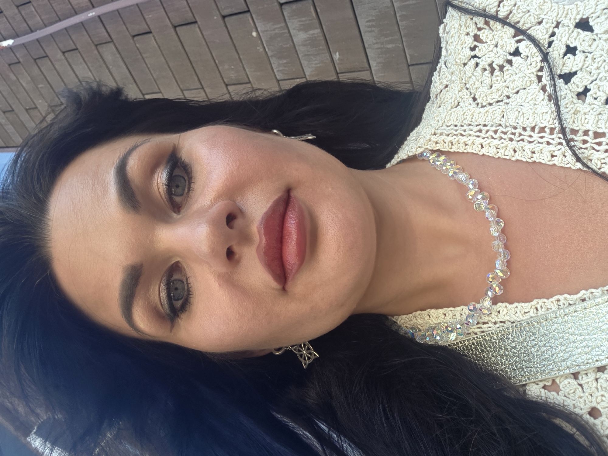 A woman with dark hair and prominent makeup poses for a selfie outdoors in front of a brick wall. She is wearing a cream-colored crocheted garment and a clear beaded necklace, with silver earrings. The background suggests an urban environment, possibly in Sochi, Russia, given the brickwork and clear sky.