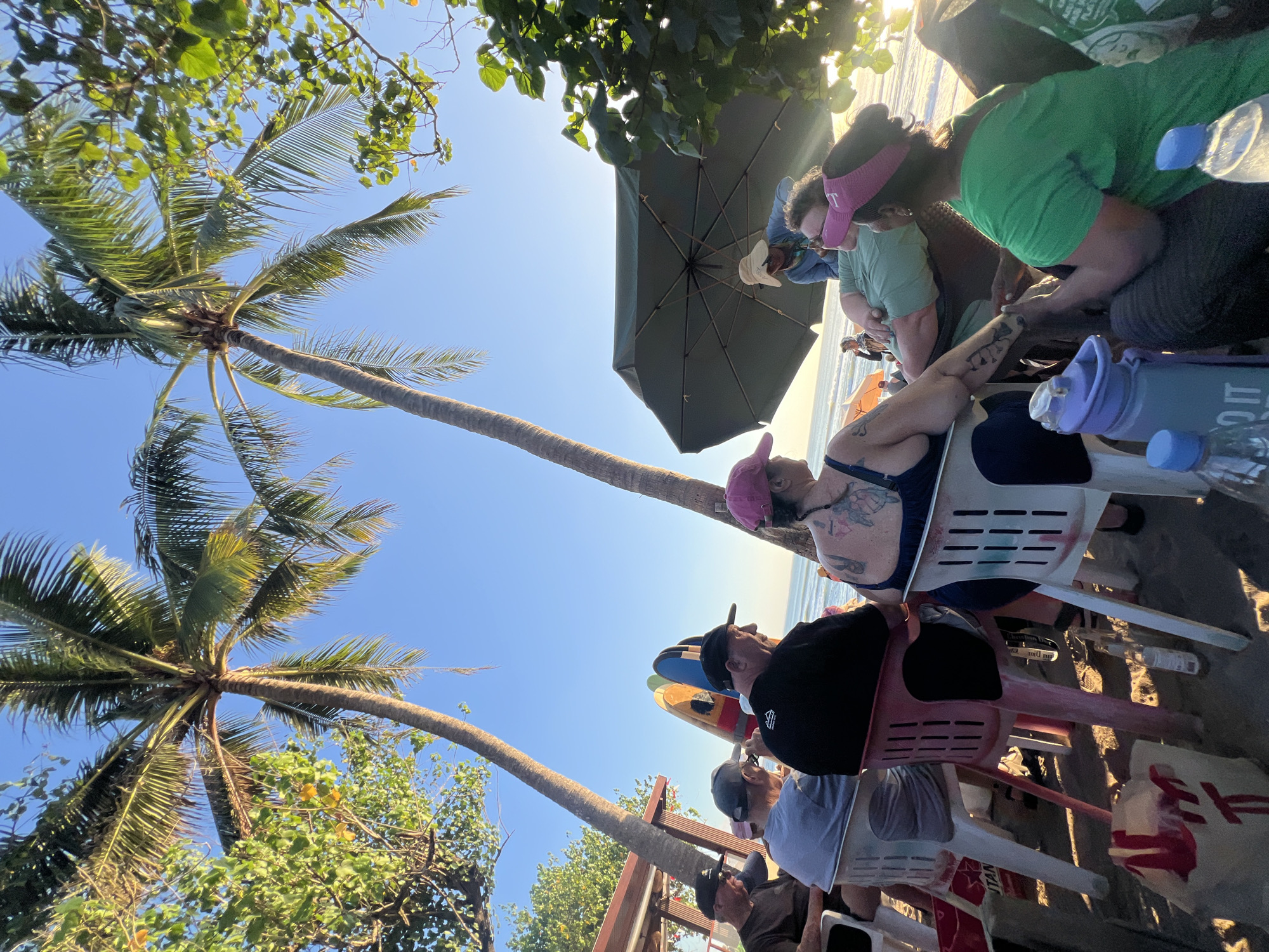 A group of people are sitting on chairs on a beach in Kuta, Indonesia, facing the ocean. There are palm trees and foliage in the foreground and background. The sky is clear and blue, and the sun is shining. Some of the people are wearing hats, and one person is wearing a visor. There are surfboards leaning against a building in the background. An umbrella is providing shade for some of the people. The overall scene suggests a relaxed, casual gathering on a tropical beach.