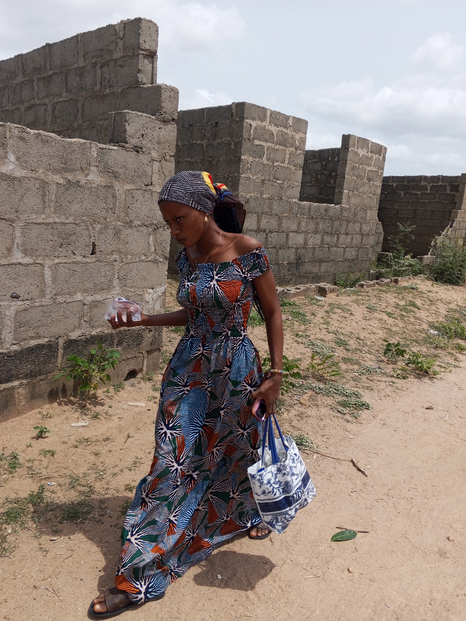 A young woman walks outdoors in Adonkolo, Nigeria, against a backdrop of unfinished concrete block structures. She has dark skin and is dressed in a vibrant, off-the-shoulder, floor-length dress adorned with a colorful geometric or floral pattern in shades of blue, orange, green, and white. Her head is covered with a black and white patterned headwrap.
In her right hand, she holds a small, clear plastic package, possibly containing money or other items. In her left hand, she carries a white and blue patterned tote bag, with a mobile phone in a pink case partially visible within or near the bag. She wears a small gold-colored earring and a beaded bracelet on her left wrist. Her gaze is directed downwards and slightly to her left, conveying a focused or pensive expression as she moves along.
The setting features sandy, dusty ground with sparse green vegetation. In the background, multiple grey concrete block walls stand unfinished, suggesting a construction site or a developing area. The sky is overcast or partly cloudy, indicating it is daytime. No discernible text is visible in the image.