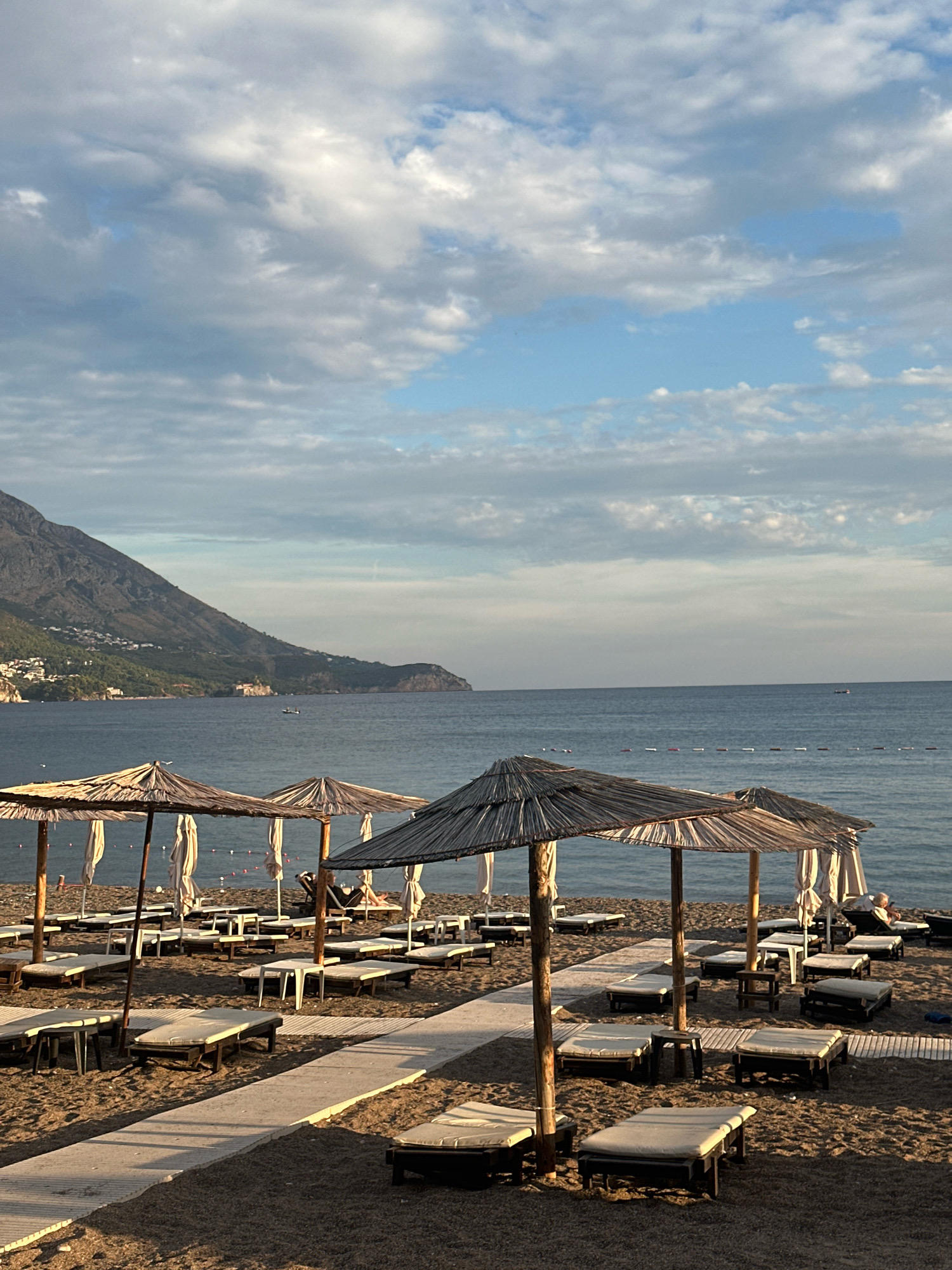 The image depicts a serene beach scene in Boreti, Montenegro, under a partially cloudy sky at what appears to be either sunrise or sunset. The foreground is dominated by rows of empty lounge chairs and straw umbrellas on a sandy beach. White wooden walkways crisscross the sand, leading towards the water. The sea is calm and blue, with a few buoys marking a designated swimming area. In the middle ground, a distant coastline is visible, featuring a verdant mountainside dotted with houses and buildings, suggesting a town or village nestled into the landscape. The sky is filled with soft, diffused clouds, some tinged with the warm hues of twilight. The overall atmosphere is peaceful and inviting, capturing the tranquility of a Mediterranean coastal setting.