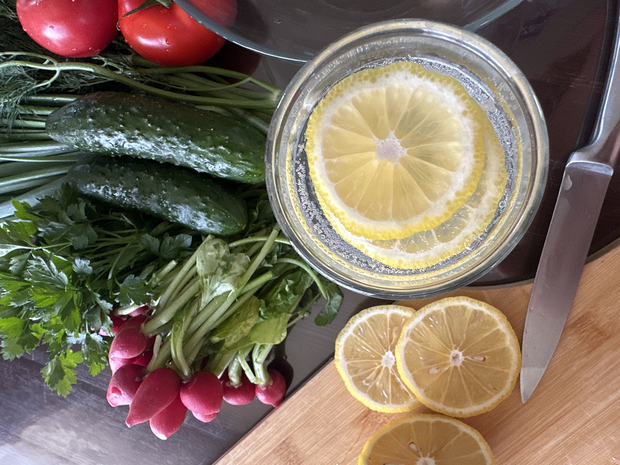 The image shows a collection of fresh vegetables and fruit arranged on a dark surface and a light-colored wooden cutting board. There are two red tomatoes, two green cucumbers, radishes, and herbs (parsley and dill). A clear glass bowl filled with sliced lemons and water is also visible. Some lemon slices are also present on the wooden cutting board. A knife is positioned on the cutting board near the lemon slices. The image suggests a preparation scene for a meal or drink. There is no evidence to indicate this image was taken in Chaikovsky, Russia.