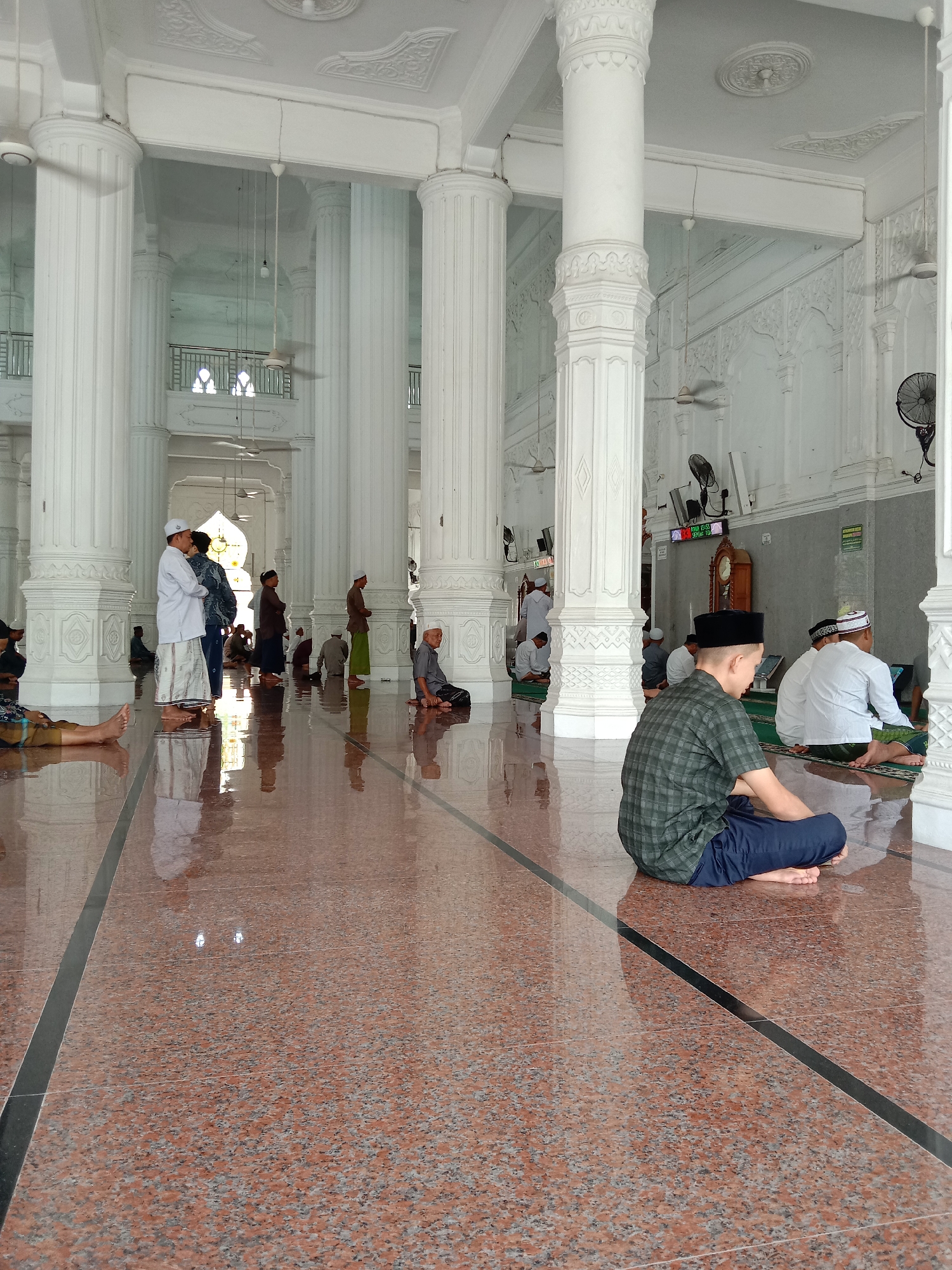 The image captures the interior of the Islamic Center Lhokseumawe mosque in Lhokseumawe, Indonesia. The scene is populated with several individuals, predominantly men, some dressed in traditional Muslim attire, including white caps (peci). Some are seated on the floor, while others are standing and engaged in conversations. One man is sitting with his legs crossed in the foreground. The mosque's architectural elements are prominent, featuring tall, fluted white columns that support the structure. The floor is a polished reddish-brown tile with black strip accents. A clock is visible on the wall in the background. Multiple ceiling fans are suspended from the ceiling. The lighting is bright.
