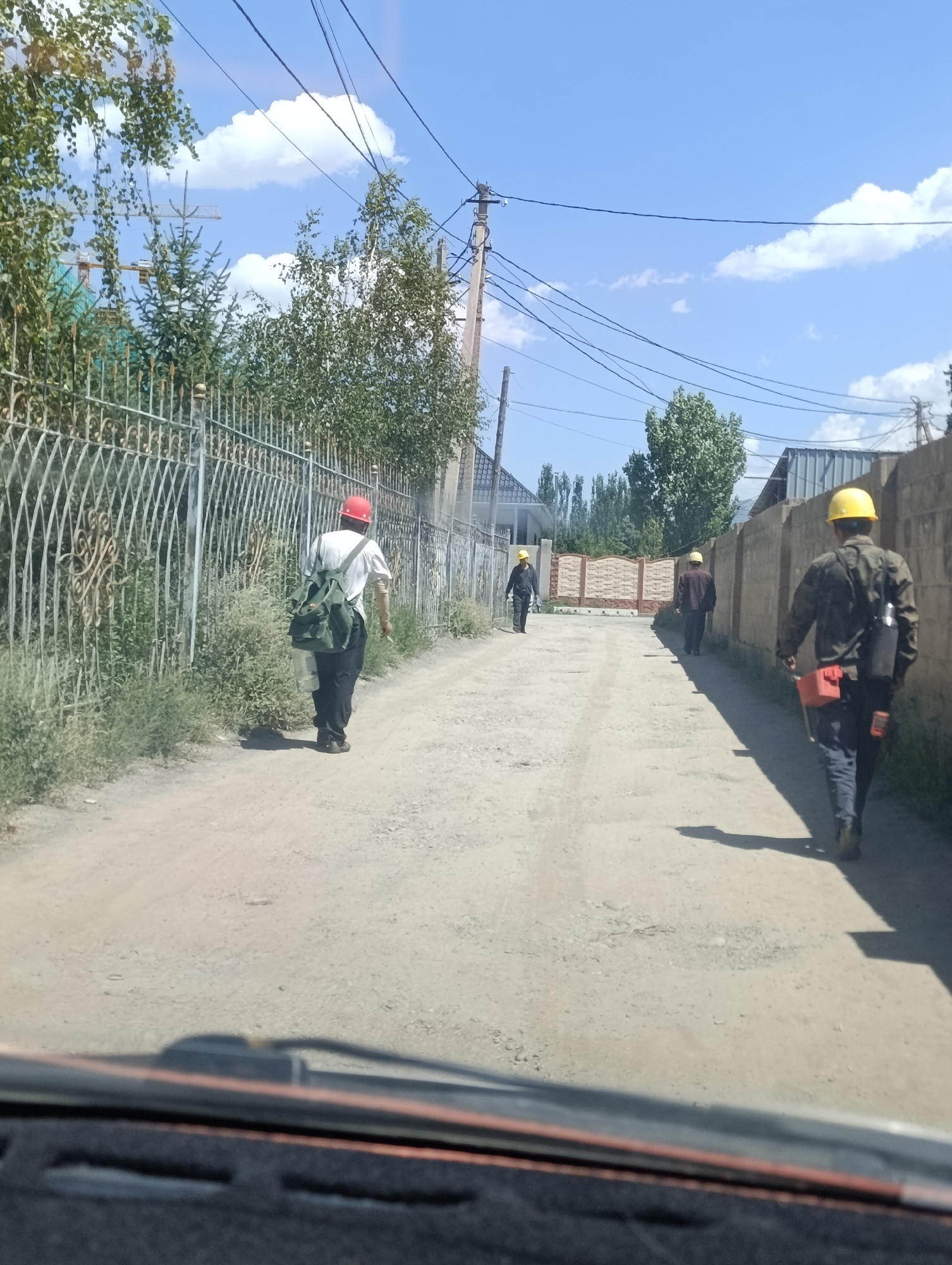In Naryn City, Kyrgyzstan, a dirt road leads through a residential area with low-rise buildings and fences. Four individuals, all wearing hard hats, are walking away from the camera. The first person, in the foreground and to the right, wears a camouflage jacket and carries a toolbox and a cylindrical container on their back. To the left and further down the road, a person in a white shirt and black pants walks with a green backpack. Two other individuals, further in the distance, are also walking along the road. The scene is set under a clear blue sky with scattered clouds.