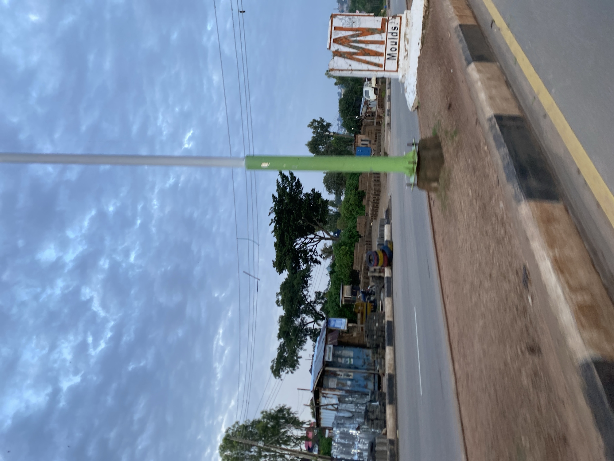 This media file depicts a roadside scene in Jos, Nigeria, likely during the late afternoon or early evening given the overcast sky. The sky is filled with dramatic, textured clouds, suggesting a possibility of rain or recent rainfall.
The primary focus is a street with a light pole prominently placed in the foreground, painted green. Adjacent to the street is a brown, dirt embankment with a curb featuring black and white markings.
On the right side of the street, a sign with a stylized "M" and the word "Moulds" is visible. Further down the road, a small stall or shop with a corrugated metal roof and blue siding is present, with various materials, possibly construction or metal supplies, stacked in front. Beyond this, more structures and trees can be seen, hinting at a bustling roadside market or commercial area.
The road itself appears to be a paved surface with a white line marking the lane. The presence of tires stacked along the roadside and a motorcycle parked near the stall suggest a local transport hub or a place where goods are sold and services are rendered. No people are clearly identifiable as the main subjects, though the scene implies a functioning community. The overall impression is one of everyday life in a busy Nigerian town.