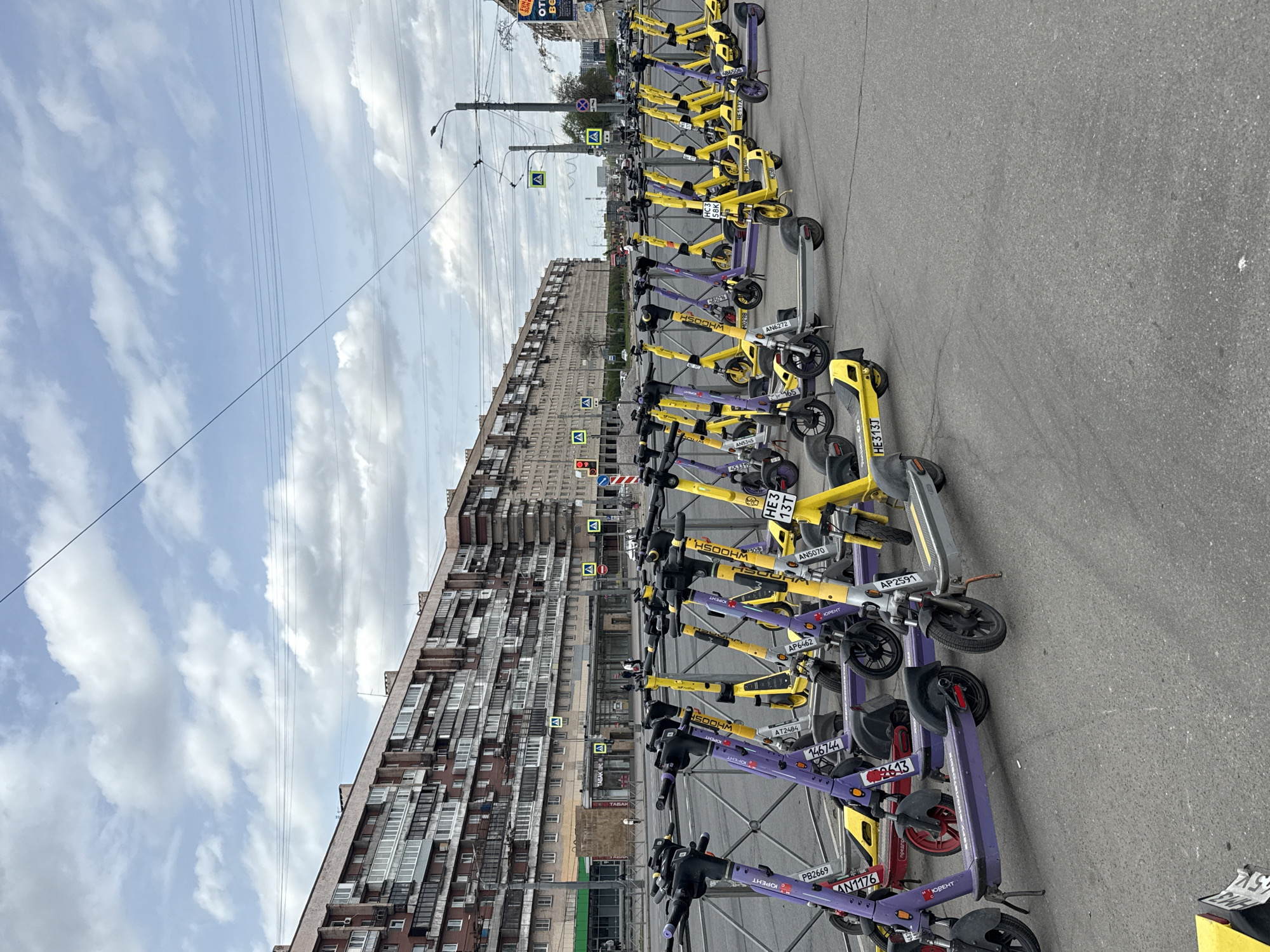 The image shows a row of electric scooters parked on an asphalt surface in Saint Petersburg, Russia. The scooters are branded "WHOOSH" and "ЮРЕНТ" and have a mix of yellow and purple colors. A tall, Soviet-era apartment building with multiple rows of windows is in the background. Street signs, lampposts, and power lines are also visible. The sky is blue with scattered white clouds.