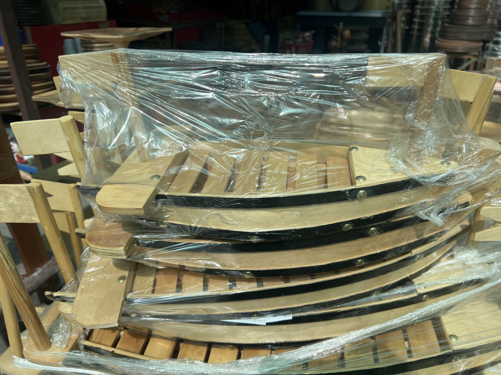 The image shows a stack of wooden boat-shaped trays, possibly for serving food, wrapped in clear plastic. The trays are made of light-colored wood with dark accents and have a curved, segmented design. They are stacked tightly together, with several layers visible. The background is blurred and appears to be an indoor setting, possibly a market or storage area, with shelves and other objects in the distance. There are no people visible in the image. The scene is brightly lit, and the plastic wrapping creates reflections and highlights on the surfaces of the trays. A label is partially visible on one of the trays at the bottom of the stack, reading "70 Enigo 40-21". The image does not provide any specific visual cues to confirm its location in Eshanguzar, Uzbekistan.