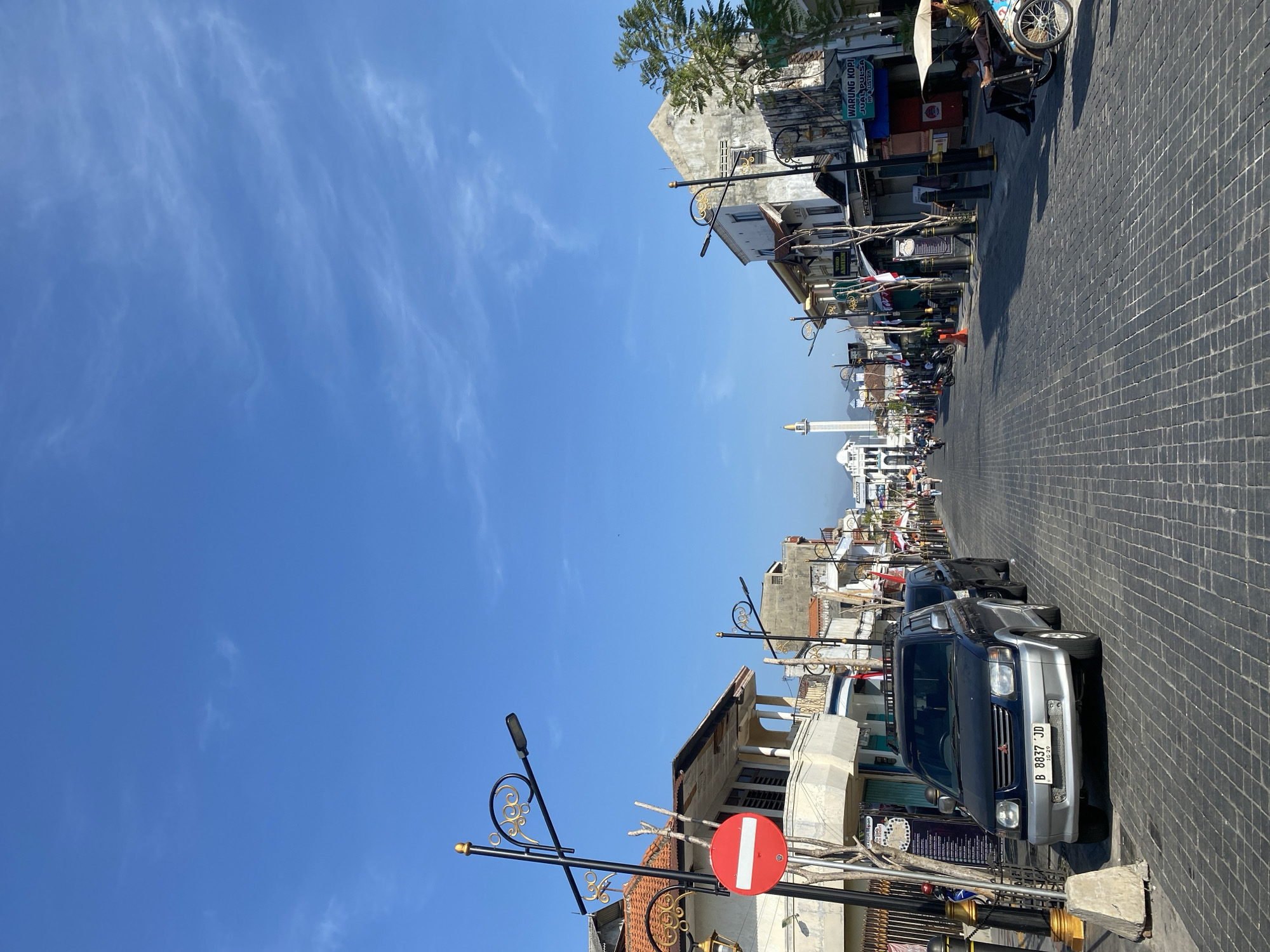 This is a street scene in the city of Kediri, Indonesia, likely on a clear, sunny day given the bright blue sky with wispy clouds. The street is paved with dark cobblestones and runs towards a distant view of a white minaret, suggesting a significant mosque or religious building. On either side of the street are low-rise buildings, many of which appear to be shops or businesses, indicated by signage such as "WARUNG KOPI JUAL PULSA HP & LISTRIK". Decorative lampposts with ornate gold detailing line the street. Several vehicles are present, including a dark SUV parked on the left with a license plate reading "B 8837 JD". A pedicab, or becak, carrying two people is visible on the right side of the street. The buildings have a mix of architectural styles, with some featuring balconies and awnings. There are a few people visible walking and sitting, and some flags are displayed along the street. The overall impression is of a busy urban street with a blend of traditional and modern elements.