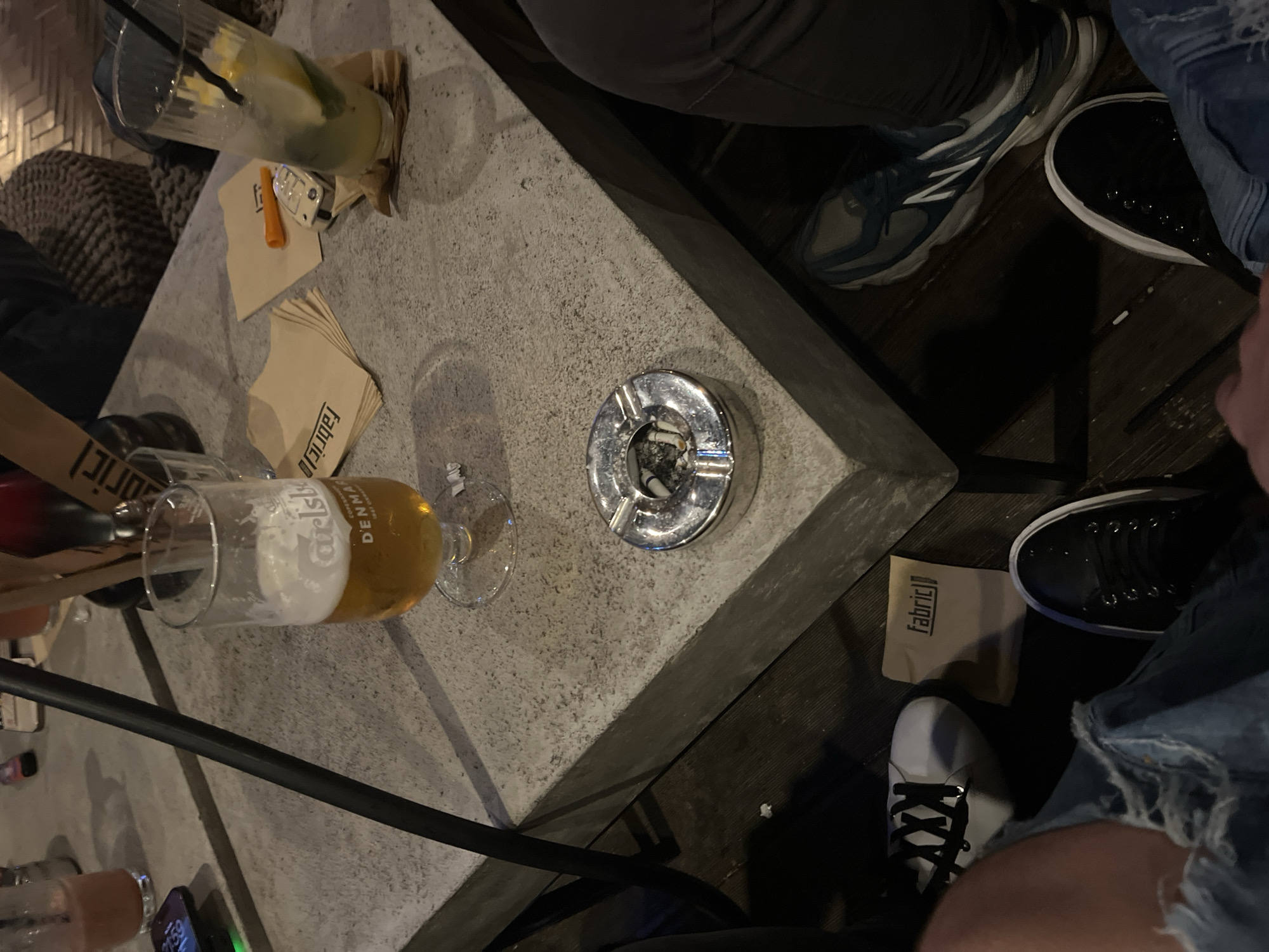 The image presents a top-down view of a casual evening gathering at what appears to be an outdoor or semi-outdoor bar or cafe in Burgas, Bulgaria. The scene centers around a sturdy, light grey concrete table, indicating a relaxed social setting.
On the table, several items are visible: a tall glass holding a cocktail with ice, citrus slices, and a straw; a half-full glass of "Carlsberg" beer, marked "DENMARK"; and a chrome ashtray containing several cigarette butts. Beige napkins, some folded and some crumpled, are scattered across the table, with visible text "Fabriclo" on a stack of clean napkins. A metallic car key fob and a small orange plastic object rest on one of the napkins. A smartphone on the table displays the time "21:59", confirming it is nighttime.
The lower sections of at least three individuals are visible around the table. One person wears dark pants and blue and white New Balance sneakers. Another is seen in black sneakers and distressed blue jeans. A third person wears white sneakers with black laces, with a small "Fabriclo" branded pouch or bag near their foot on the dark wooden floor. Part of a dark, woven ottoman or piece of furniture is visible in the background. The overall atmosphere suggests a casual social event with drinks among friends.