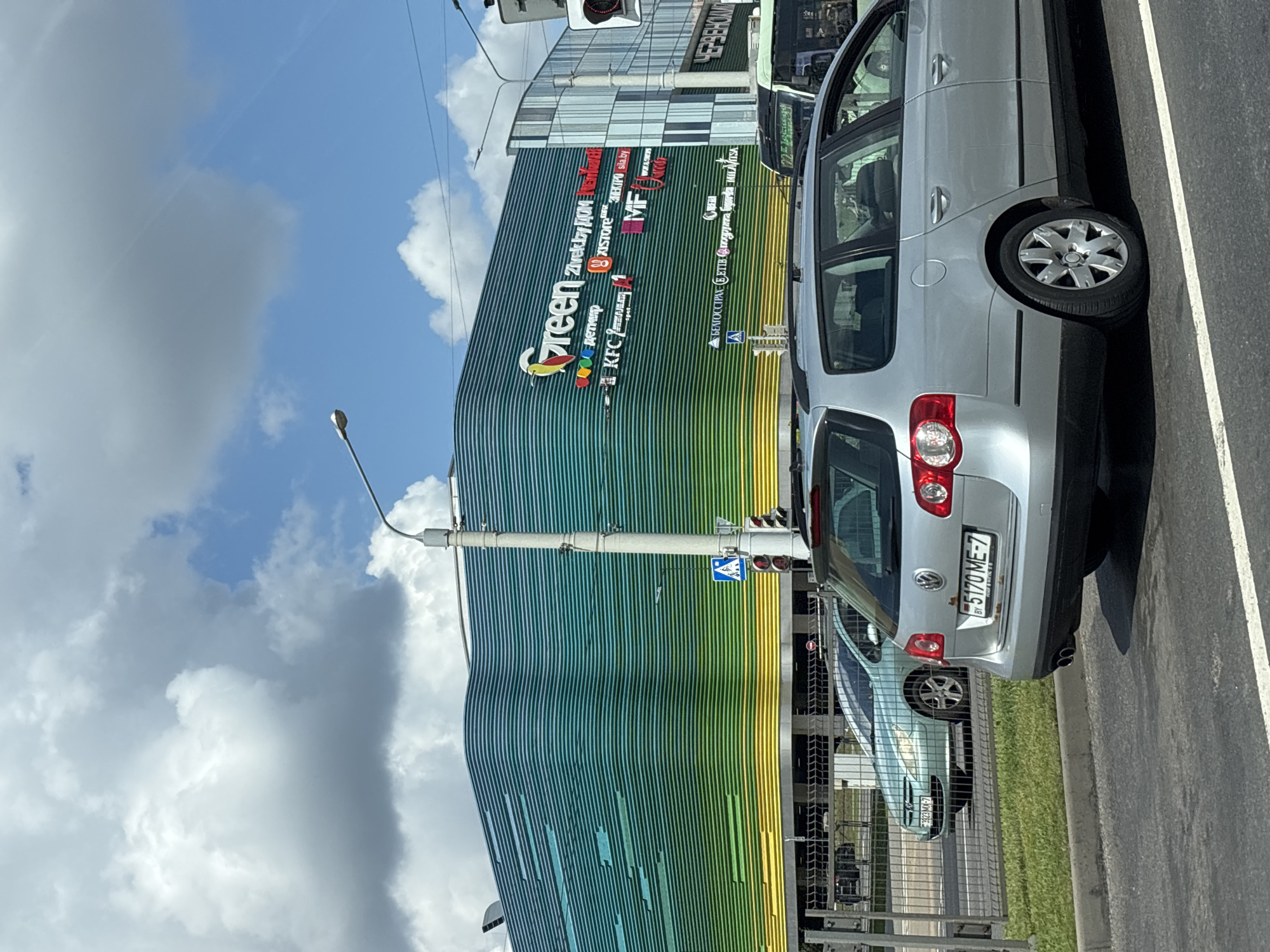 In Minsk, Belarus, a daylight street scene is captured, focusing on the "Green" shopping center, a large, modern building with a distinctive facade of horizontal wavy bands in shades of green and yellow. Various logos are visible on the building's exterior, including "Green," "Zivek.by," "KFC," and "MF." In the foreground, a silver Volkswagen Passat station wagon is parked, its rear license plate reading "BY 5170 ME-7." To the left of the Passat, a light blue Peugeot 307 hatchback is visible, its license plate partially obscured. The cars are on a paved road, separated from a sidewalk by a metal fence. A pedestrian crossing signal with red lights is mounted on a pole. Beyond the shopping center, a public bus with signage in Cyrillic script is also present. The sky above is partly cloudy with a bright blue sky peeking through.