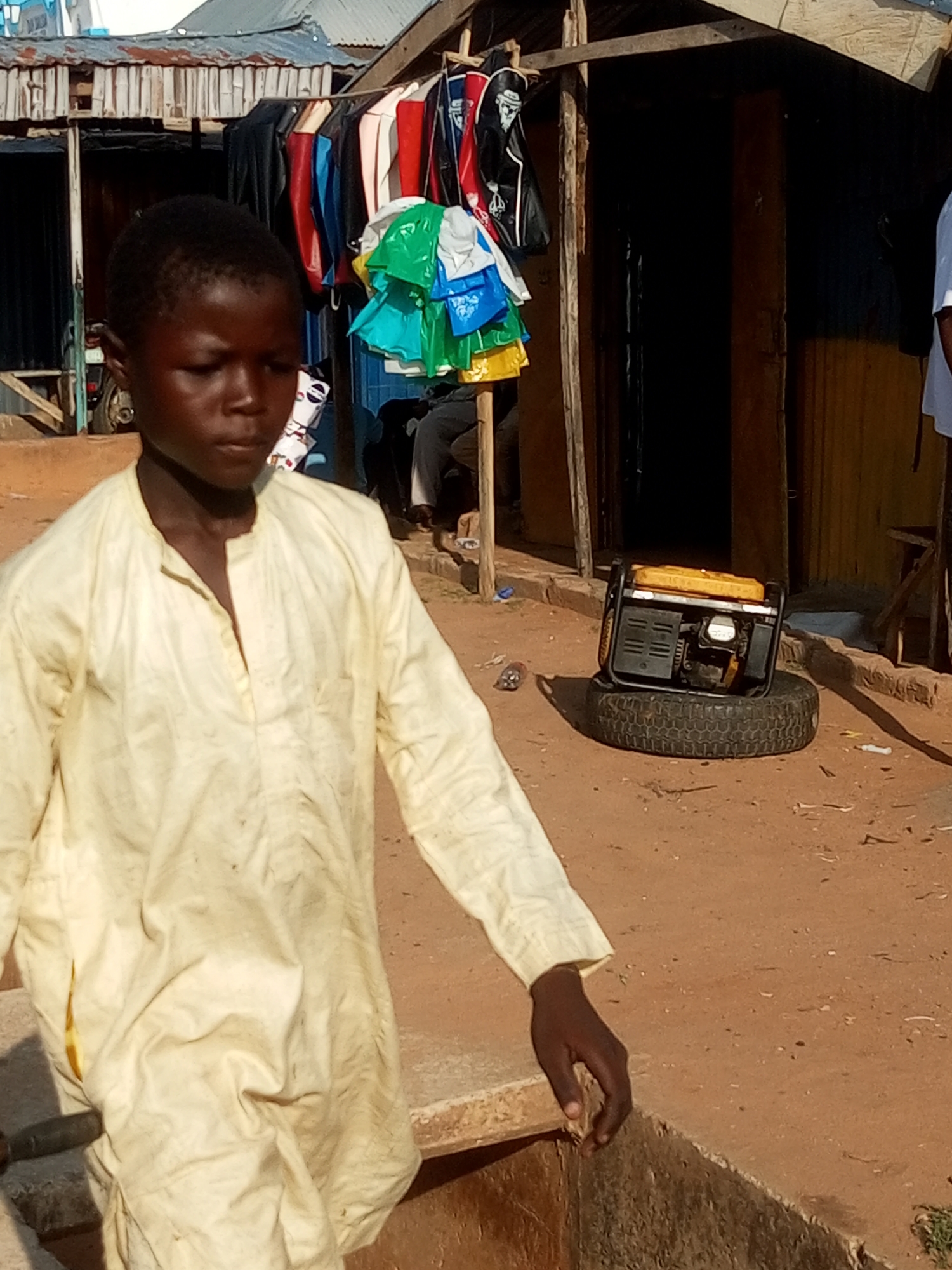 In a dusty outdoor market in Gombe, Nigeria, a young boy in a light yellow garment walks past a stall. The stall displays a rack of colorful clothing and various plastic bags in bright green, blue, and yellow. Behind the stall, a dark doorway suggests the interior of a shop or dwelling. To the right of the boy, a generator sits on a tire in the dirt. The scene is illuminated by natural light, indicating it is daytime, with the sun casting shadows on the ground. The ground itself is a reddish-brown dirt, common in arid or semi-arid regions. The overall atmosphere suggests a typical day in a bustling local marketplace, with commerce and everyday life taking place. The boy's expression appears neutral as he moves through the scene.