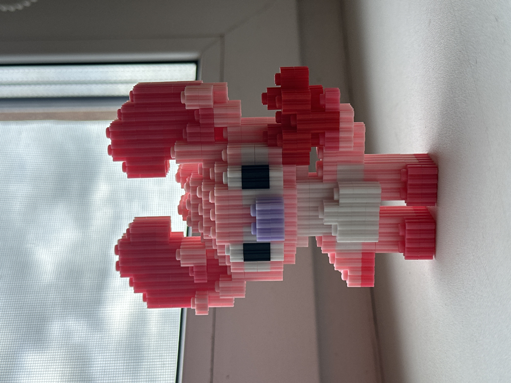 A pink, blocky figurine resembling the character Angel from Lilo & Stitch stands on a white surface in front of a window with a sheer curtain. The figurine has large ears, black eyes, a purple nose, and is holding a red heart shape in its right hand. The background shows a cloudy sky through the window. The scene appears to be indoors, on a windowsill. The location is Ждановіцкі сельскі Савет, Belarus.