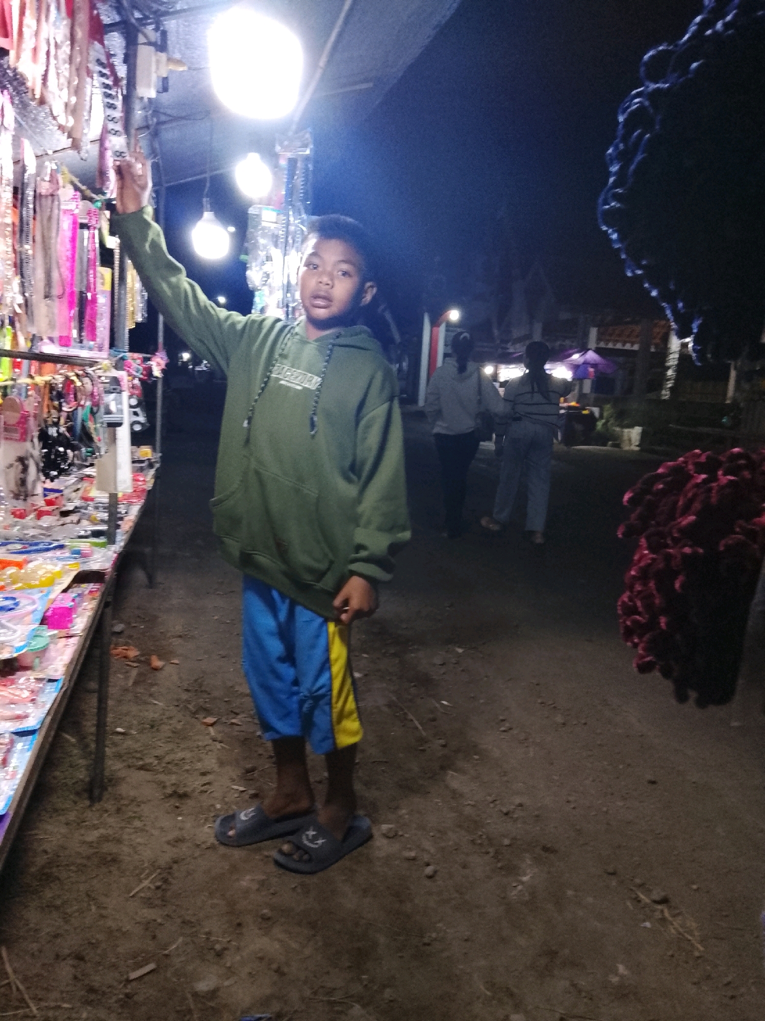 A young boy stands in front of a market stall at night. The stall displays various items, possibly accessories or small goods, illuminated by bright overhead lights. The boy is wearing a green hooded sweatshirt and blue shorts with a yellow stripe. He is looking towards the camera with his right arm raised. In the background, two women are walking away from the stall, and a large cluster of red, fluffy objects is visible on the right side of the frame. The ground is dirt, and the overall scene suggests an outdoor night market in a rural or semi-urban setting. The location is identified as Sukorejo, Indonesia.