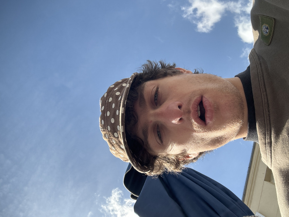 A low-angle, close-up photograph depicts a young adult male, identified as being in Vincennes, France, under a clear blue sky with scattered white cumulus clouds. The subject, positioned centrally, has brown curly hair and light stubble. He wears a brown cap with white polka dots, a black turtleneck, and a brown crewneck sweatshirt. A square, olive-green patch with a circular emblem featuring a light-colored cartoonish face is affixed to the left shoulder of his sweatshirt. His blue eyes are directed towards the camera, and his mouth is slightly open. To the left, a dark blue fabric object, possibly an umbrella or awning, is partially visible. A light-colored building facade is partially discernible in the lower-left background. The scene is brightly lit by natural sunlight.