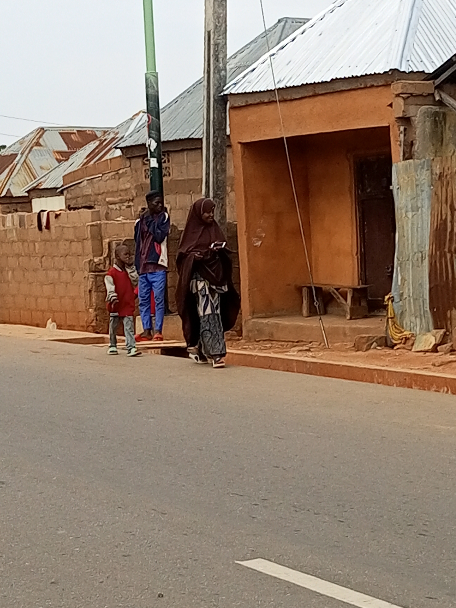 The image shows a street scene in Tumfure, Nigeria. A young boy wearing a red jacket and grey pants stands on the side of the road, facing left. Behind him, a young man in a blue and white jacket and blue pants stands next to a wall. To the right of the young man, a woman in a brown hijab and a patterned dress walks forward, looking down at a phone in her hands. A light green lamppost stands near the young man. The buildings in the background have corrugated metal roofs and are constructed from brick. The street is paved with asphalt and has a white line marking its edge.