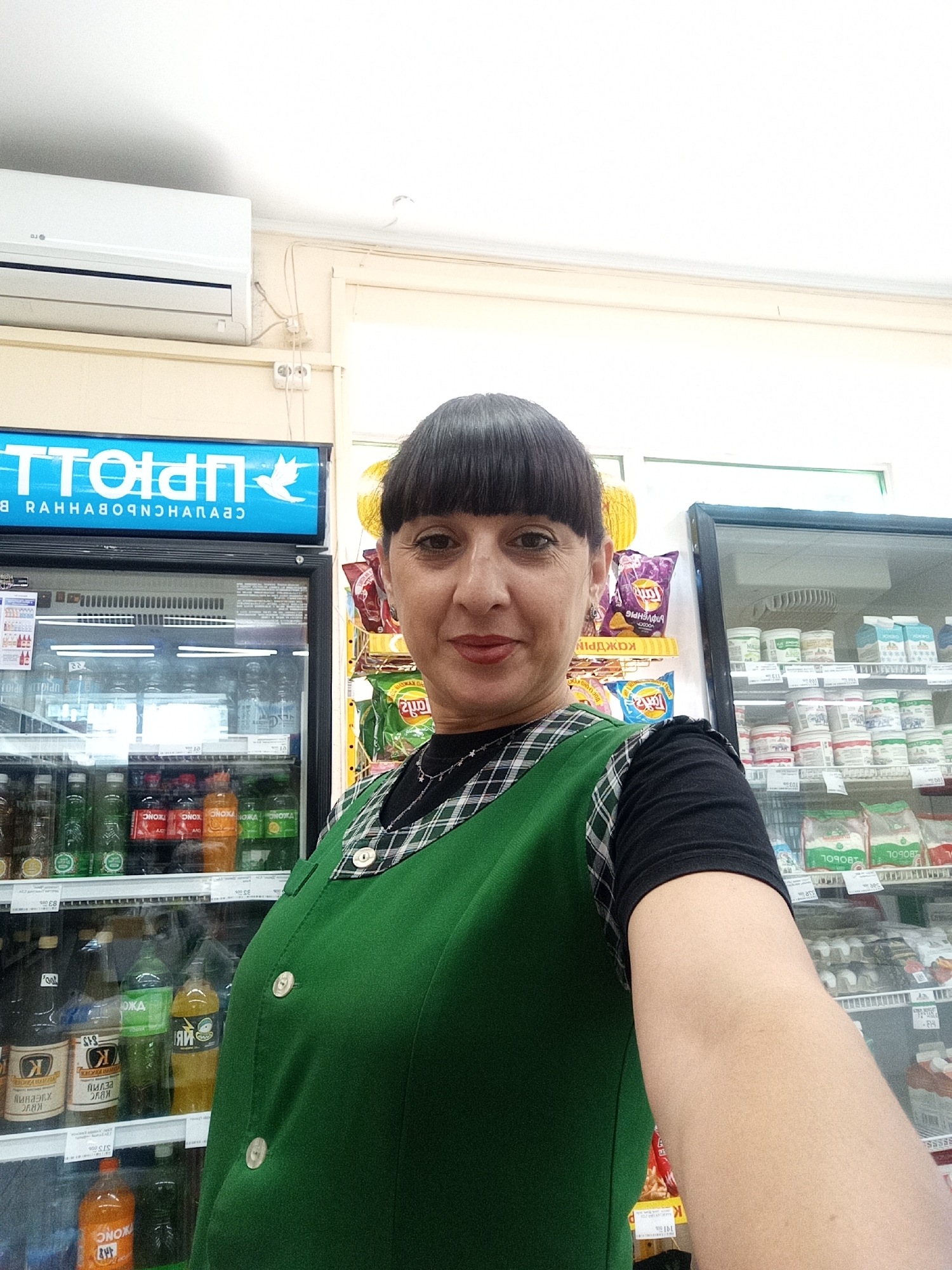 In the city of Belogorsk, Russia, a woman with dark hair, bangs, and bangs stands in front of a refrigerated display case in a store. She is wearing a green vest over a black shirt and a silver necklace. The display case contains various bottled drinks. Behind her, shelves are stocked with snacks and other products. An air conditioning unit is mounted on the wall above the display case. The lighting is bright, typical of a retail environment.