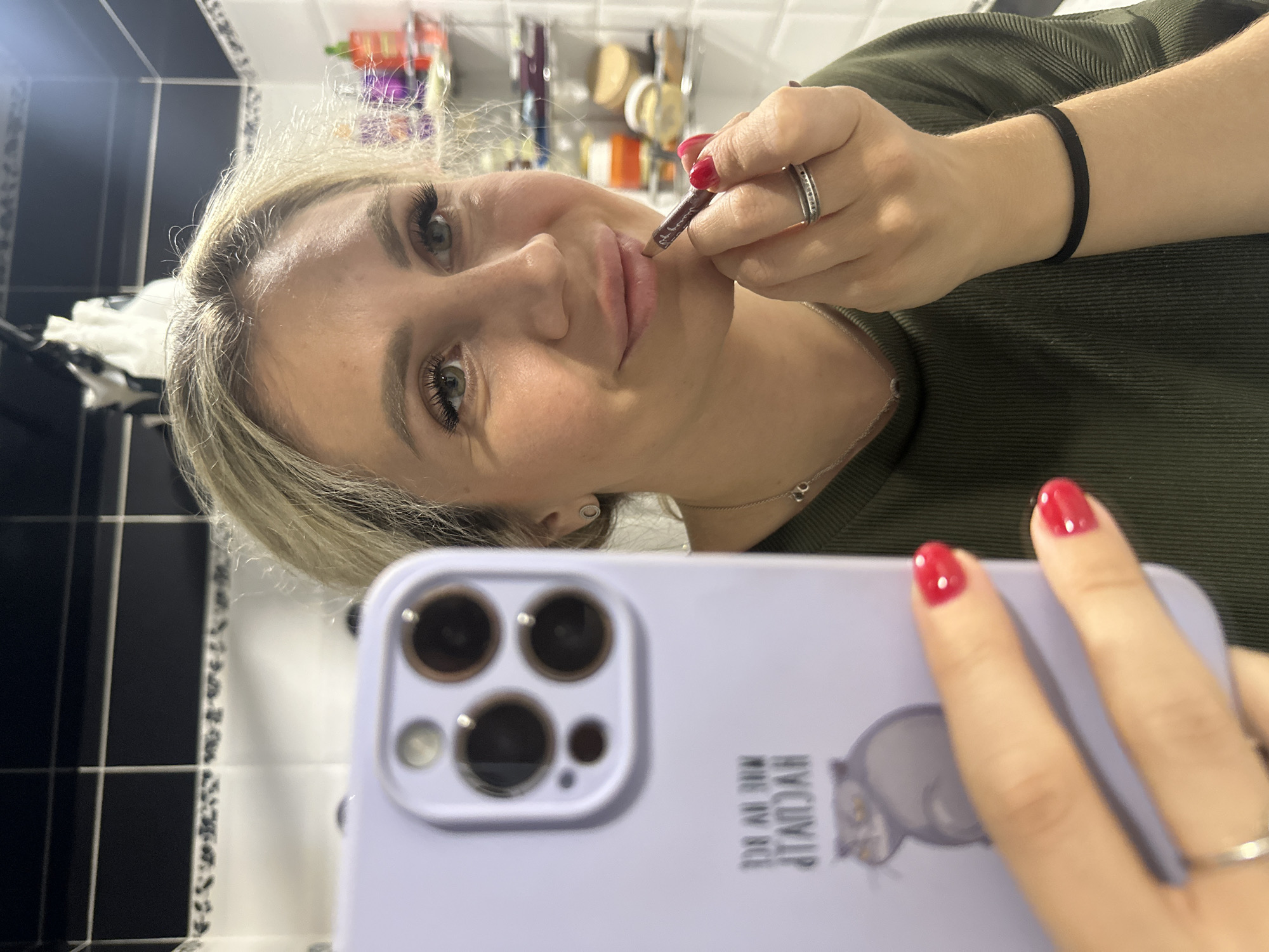 A blonde woman with full lips and dramatic eyelashes is applying lip liner to her lips. She is holding a purple smartphone with a cat sticker on its case in her left hand, which is reflected in a mirror. The woman is wearing a dark green, ribbed, long-sleeved shirt and several rings on her fingers, including a wedding band. Her fingernails are painted red. The background appears to be a bathroom, with black and white tiled walls and shelves filled with various toiletries and bottles. The lighting is somewhat dim, creating shadows. The woman is looking into the mirror as she applies makeup. There are no visible indicators of the city of Novoaltaysk, Russia, in the image.