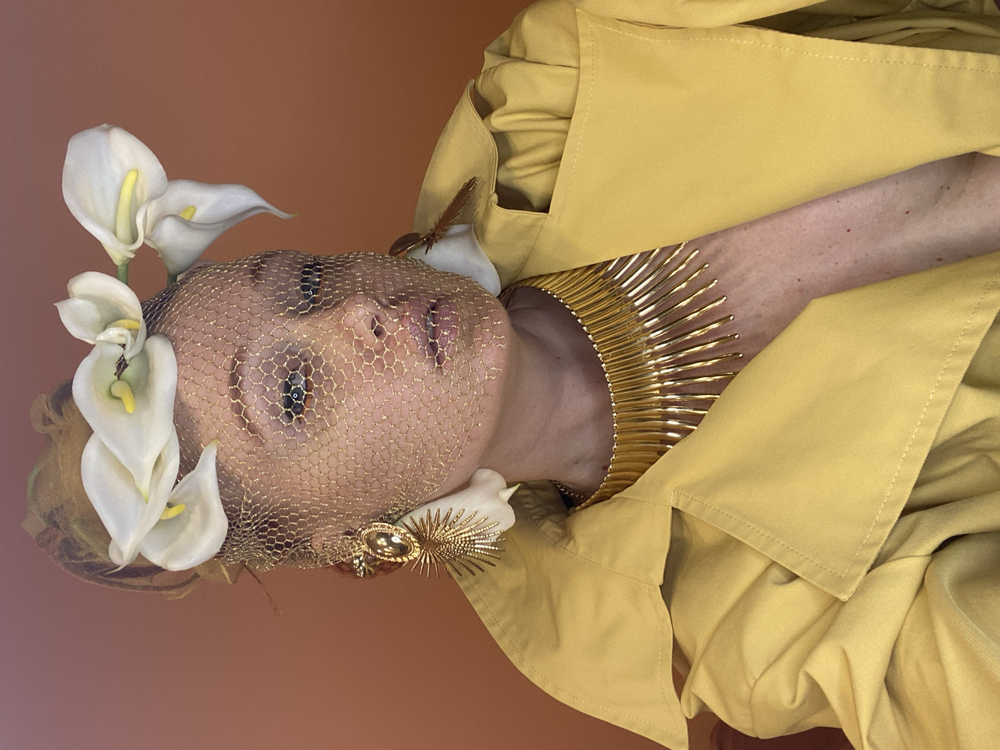 This is a close-up, vertical, eye-level shot of a person adorned with floral and metallic accessories. The subject is wearing a yellow garment that appears to be a trench coat, with its collar and lapels prominent. A delicate gold mesh veils the subject's face, obscuring most of it, but revealing one eye with dark makeup. White calla lilies are arranged around the subject's head, interspersed with what looks like a small brown feather or decorative element. A striking gold collar, resembling a sunburst with radiating spikes, encircles the subject's neck. A gold brooch, also sun-shaped, is attached to the mesh veil near the subject's cheek. The background is a solid, warm brown color. The lighting is soft and diffused, casting subtle shadows and highlighting the textures of the fabric and accessories. The image evokes a sense of artistic or fashion-oriented portraiture, with a slightly surreal or theatrical quality due to the elaborate headwear and veil. There is no visible text in the image. The setting does not provide specific cues about the time of day, weather, or location beyond what might be inferred from the attire, which is not particularly indicative of a specific climate or time. The emotion conveyed is one of artistic expression and perhaps a touch of mystery due to the veiled face.