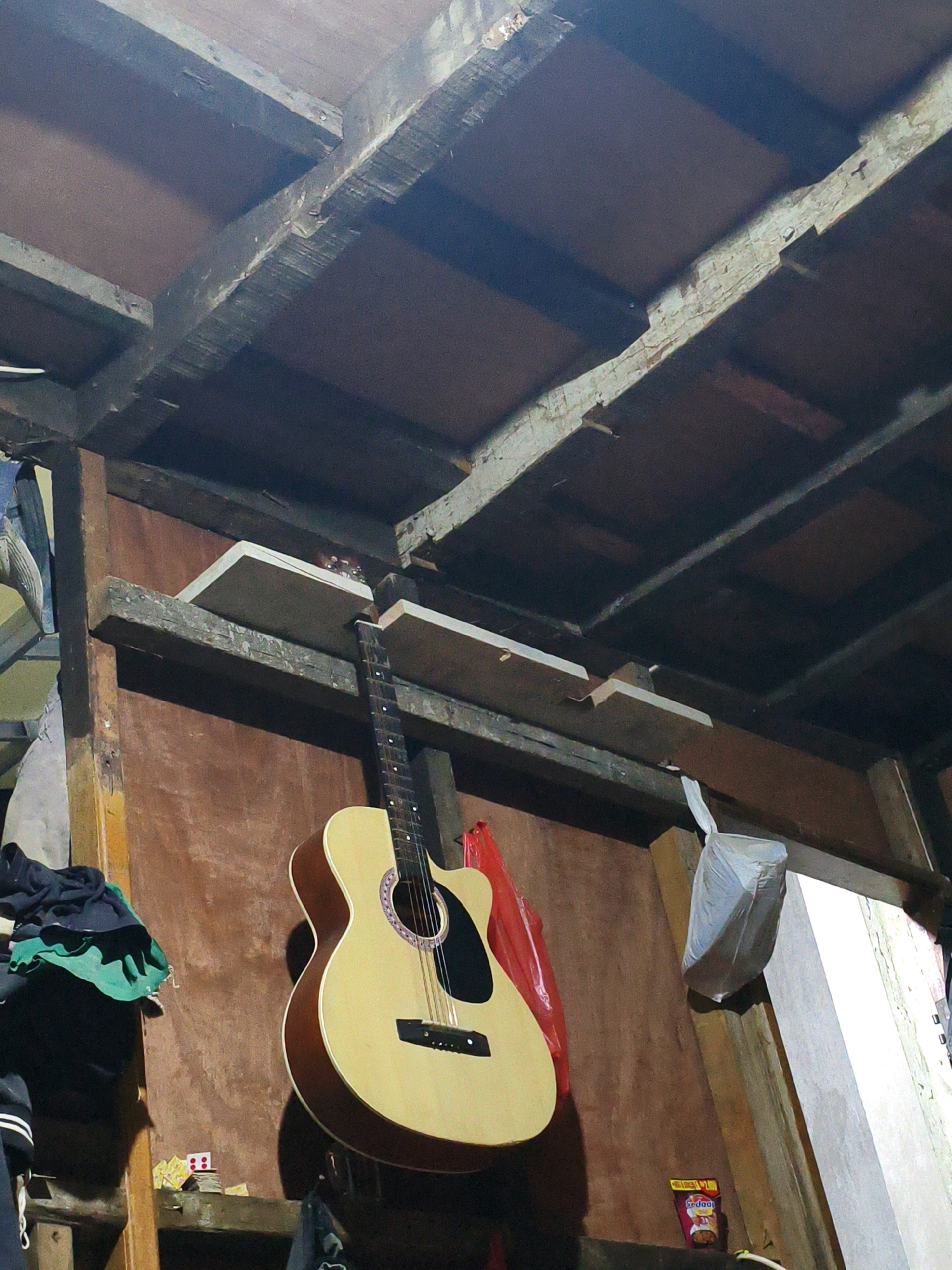 The image depicts a dimly lit interior scene, likely a workshop or storage area, with an acoustic guitar prominently displayed. The guitar, made of light-colored wood with a darker brown trim, hangs on a wooden wall. Above the guitar, there are exposed wooden beams and what appear to be makeshift shelves. To the left of the guitar, a pile of dark clothing, including a green fabric, is visible. To the right, a white plastic bag is suspended from a beam, and a small red bag is partially visible behind the guitar. In the lower right corner, a small packet of food, possibly instant noodles, is on a shelf. The environment suggests a rustic and utilitarian space. The image is a photograph taken in Bekasi, Indonesia.