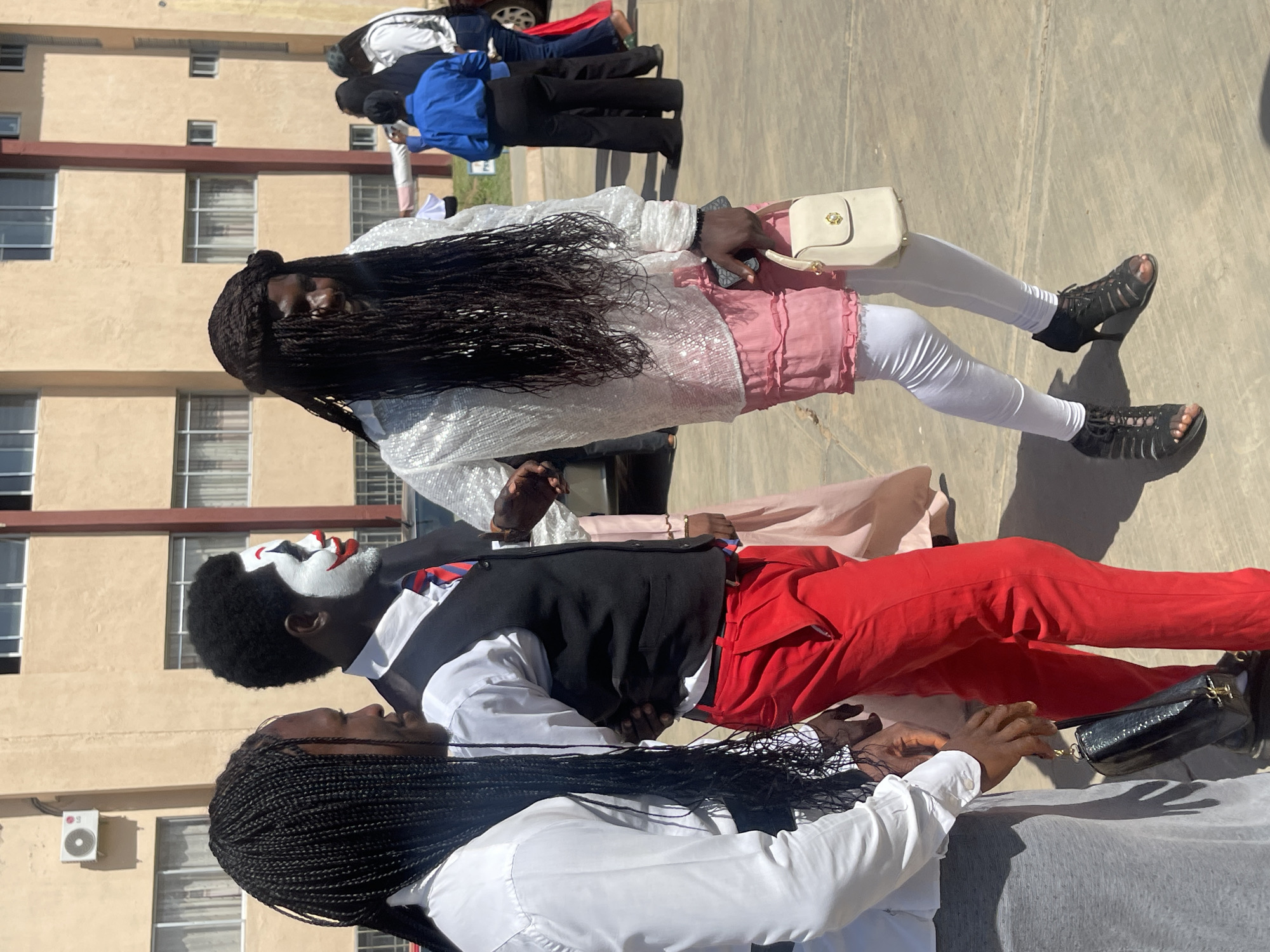 In Rafin Par, Nigeria, three individuals are captured in a medium shot outdoors, with a modern building in the background. The central figure, with clown-like face paint, wears a white shirt, black vest, red tie, and vibrant red pants, their right arm linked with the person to their left. The person on the left, with long black braids, sports a white shirt and a grey skirt, holding a black clutch bag. To the right of the clown, another individual with long dark hair cascading over their shoulders wears a shimmering white top, a pink ruffled skirt, white leggings, and black heeled sandals, carrying a small cream handbag. Several other individuals are visible in the background, with one wearing a blue shirt and black pants. The ground is paved, and sunlight casts shadows.