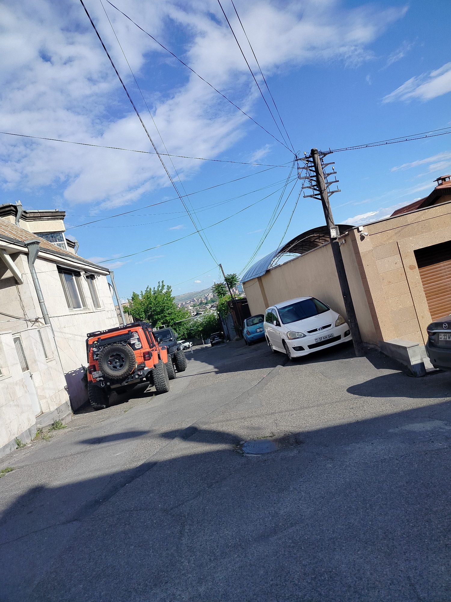 The image depicts a residential street scene in Yerevan, Armenia on a sunny day with a blue sky and scattered clouds. The street slopes downward, lined with buildings and parked vehicles. On the left side of the frame, there is a light-colored, two-story building with visible windows and drainpipes. Parked in front of this building is an orange Jeep Wrangler with a visible spare tire mounted on the back. Behind it, another dark-colored Jeep is partially visible.
On the right side of the street, a beige stone wall runs alongside the road, and a wooden utility pole stands next to it, with numerous wires crisscrossing above. A white car is parked close to the wall, with a visible license plate. Further down the street, a small blue car can be seen, as well as a view of the city beyond, hinting at Yerevan's hilly topography.
The street itself is paved with asphalt and is partly covered in shadows, adding contrast to the scene. There are no people visible in the photograph, and the focus is primarily on the built environment and parked vehicles. A portion of another vehicle can be seen parked along the wall closer to the viewer on the right.