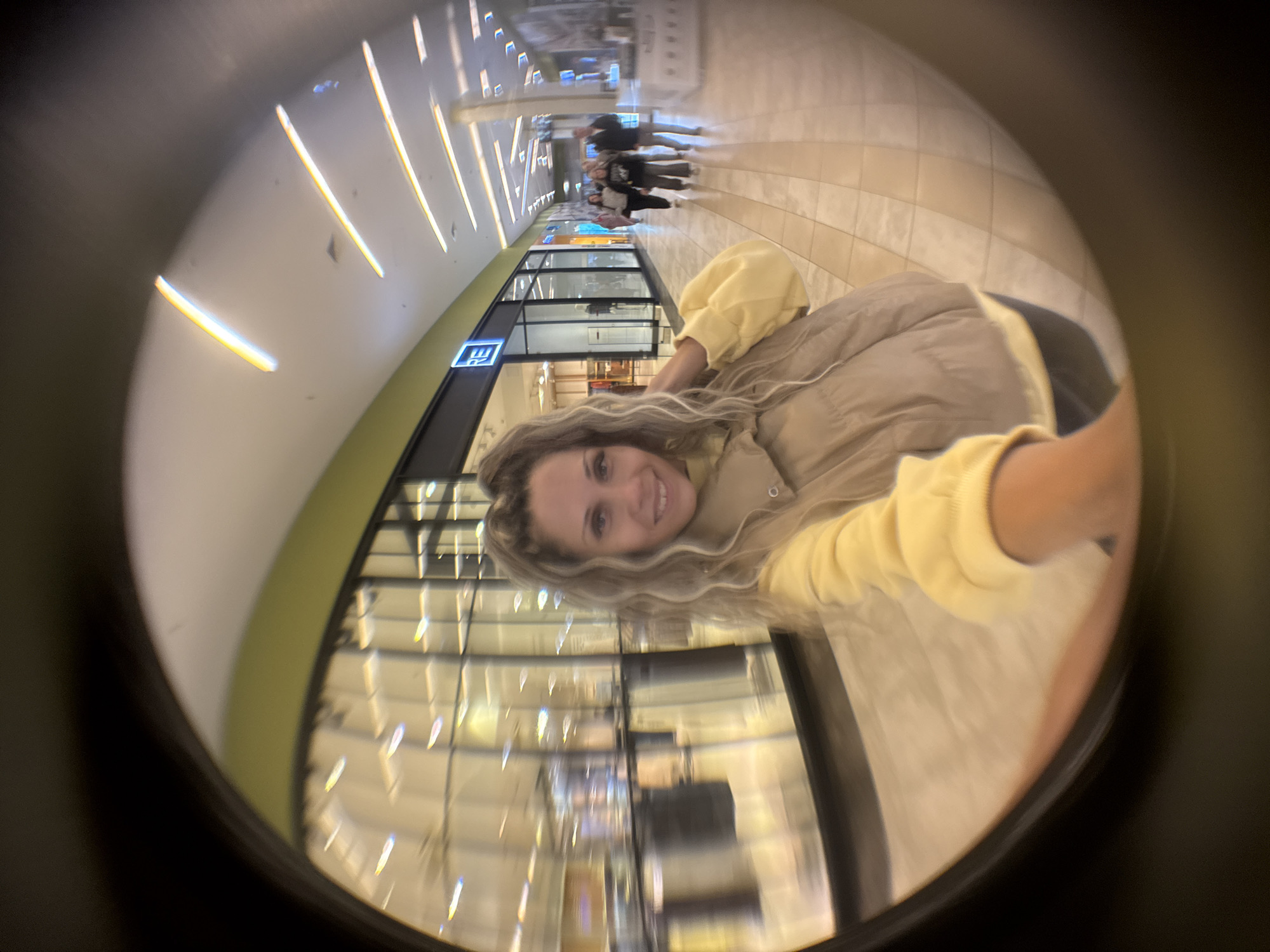 The image shows a fisheye lens selfie of a woman in a shopping mall. She has blonde, wavy hair and is wearing a light yellow sweatshirt and a beige puffer vest. She is smiling and touching her hair with her right hand. In the background, other shoppers are walking down the mall hallway, which has storefronts and illuminated ceiling lights. The mall appears to be a modern indoor shopping center with tiled floors and glass storefronts. There is a sign above one of the storefronts that reads "RE" in blue letters. The overall lighting is bright, suggesting daytime. The image does not contain any specific visual cues that would indicate its location within the city of Yaroslavl, Russia.