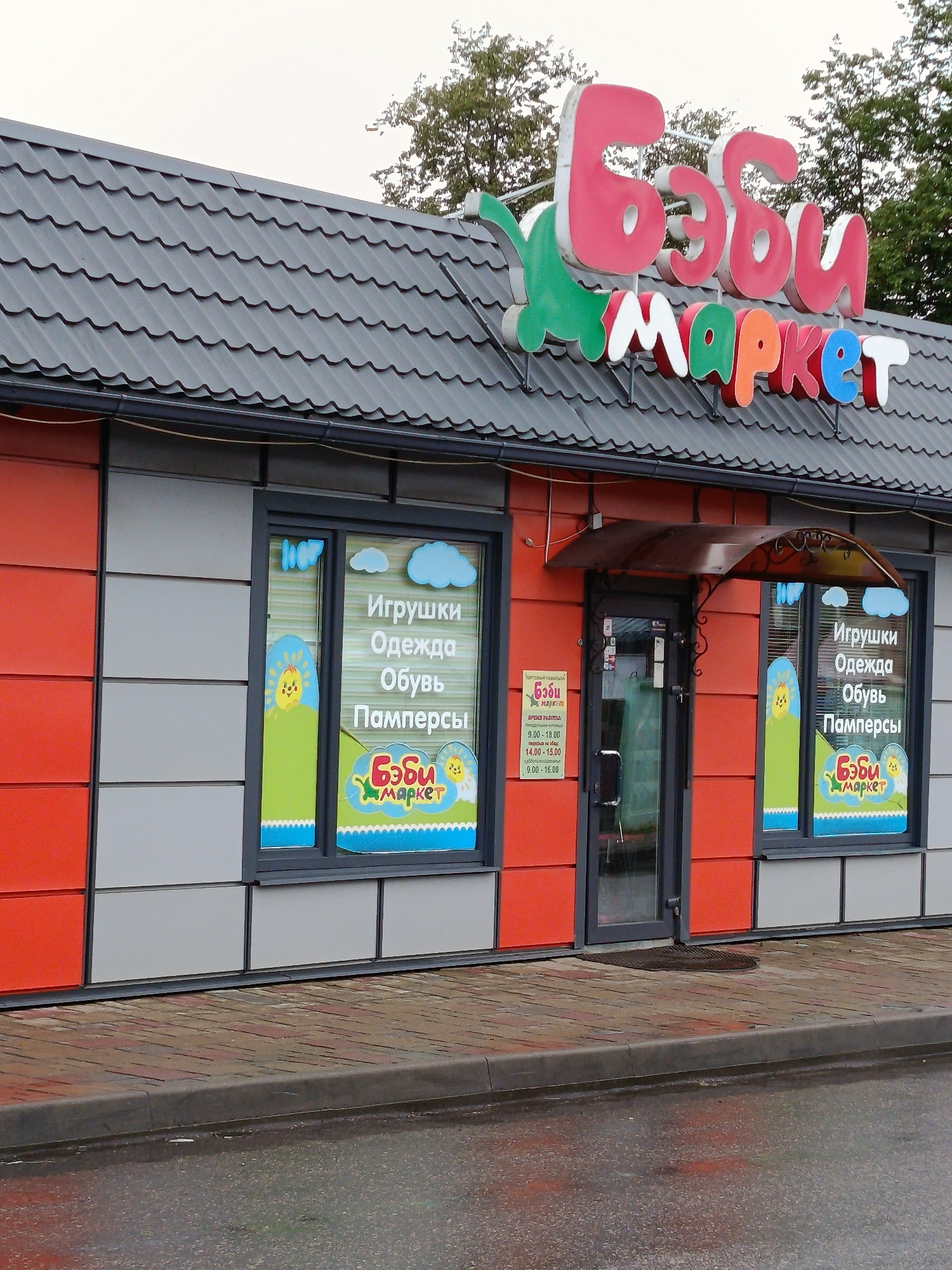 The image shows the exterior of a store called "Baby Market" located in Smalyavichy rural council, Belarus. The store has a modern facade with red and grey panels. A prominent sign with pink and colorful letters spells out the store's name. The store windows display graphics advertising toys, clothes, shoes, and diapers. There is a glass door with an awning above it. The pavement in front of the store is wet, suggesting it has recently rained. Trees are visible in the background. The scene is devoid of people.