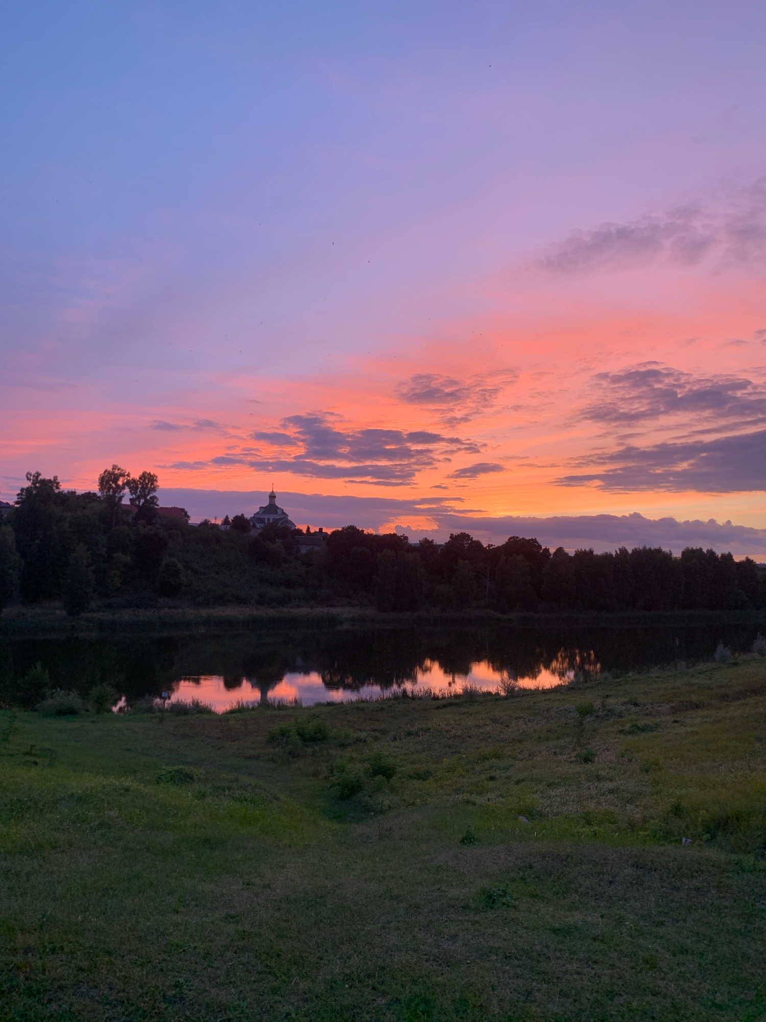 The image depicts a serene sunset scene over a body of water, with a densely wooded hill in the middle ground and a grassy foreground. In the distance, atop the hill, a white building with a green dome, likely a church, is visible, characteristic of the architecture found in the city of Kapyĺ, Belarus. The sky is painted with vibrant hues of purple, pink, and orange, with scattered clouds adding texture to the twilight atmosphere. The water reflects the colors of the sky and the silhouettes of the trees and the building, creating a mirror-like effect. The overall impression is one of tranquility and natural beauty.