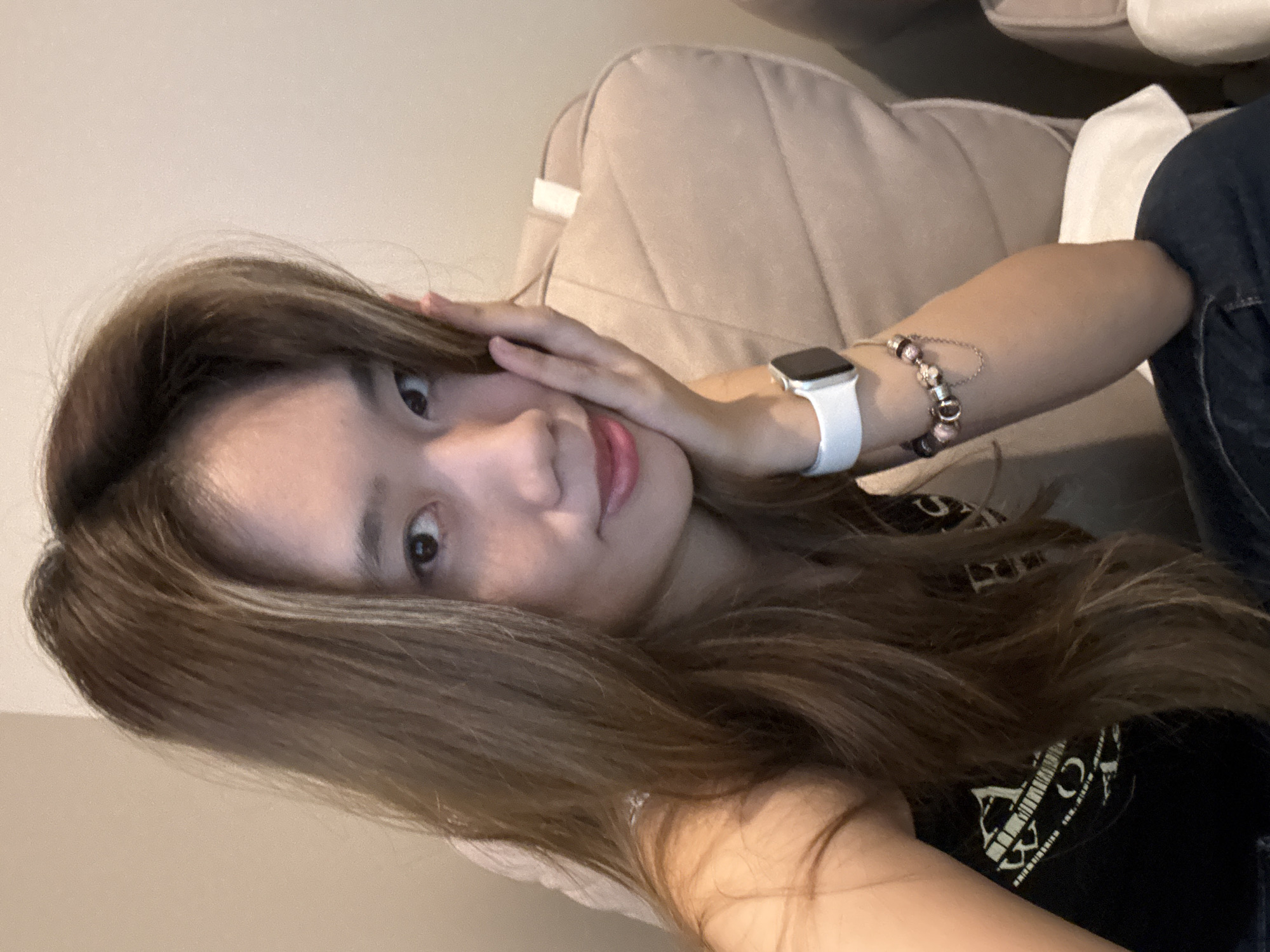 A young woman with light brown hair, wearing a black shirt, poses indoors. She has a white smartwatch and a beaded bracelet on her left wrist. Her left hand is touching her face. She is sitting or reclining on a beige upholstered surface. The location in the city of Liangjiang, China is unknown.